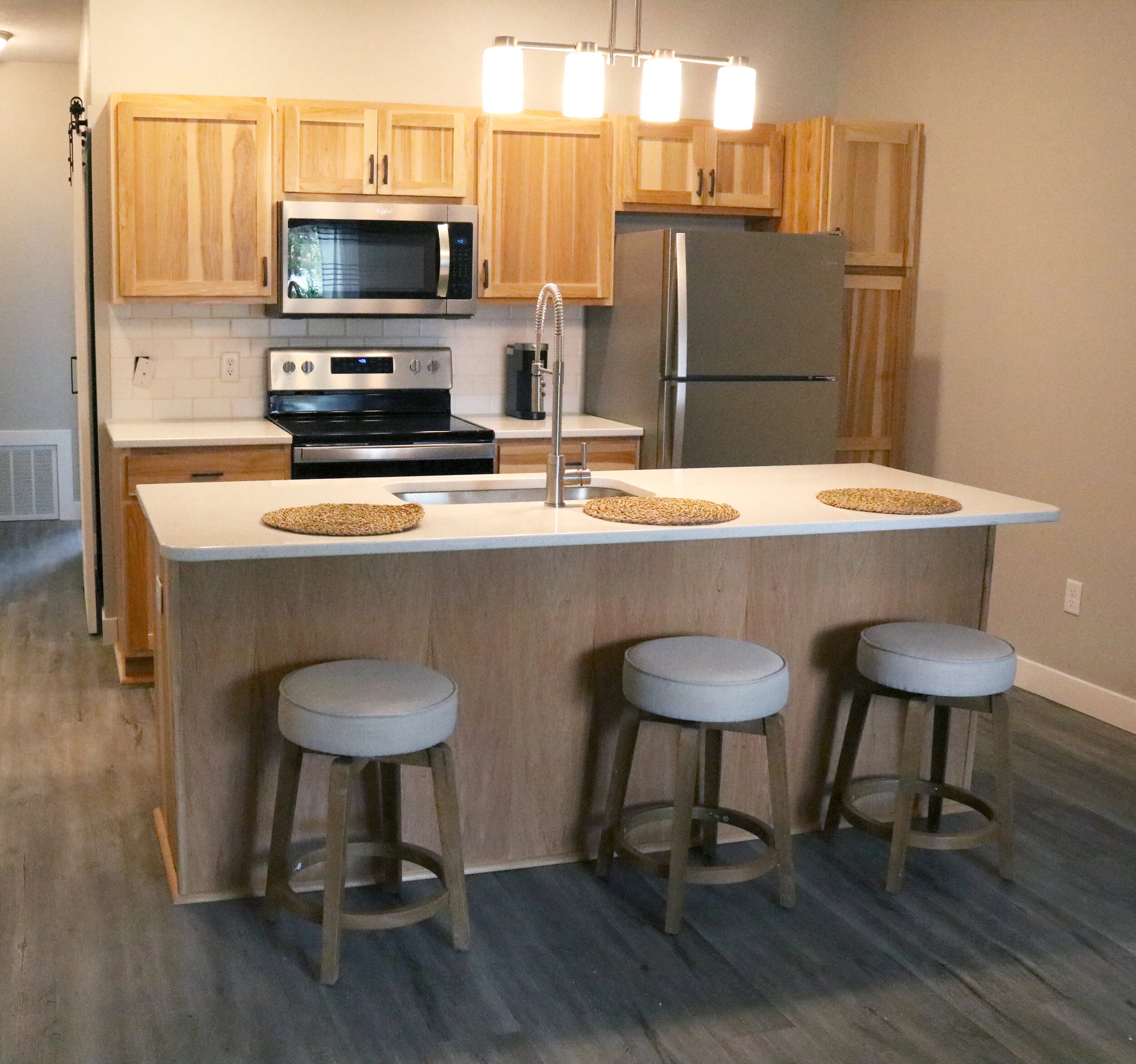 Modern kitchen with wooden cabinets, stainless steel appliances, and an island with stools. The island has a white countertop with woven placemats, a sink, and a chrome faucet. There is a stainless steel refrigerator and microwave, and overhead light