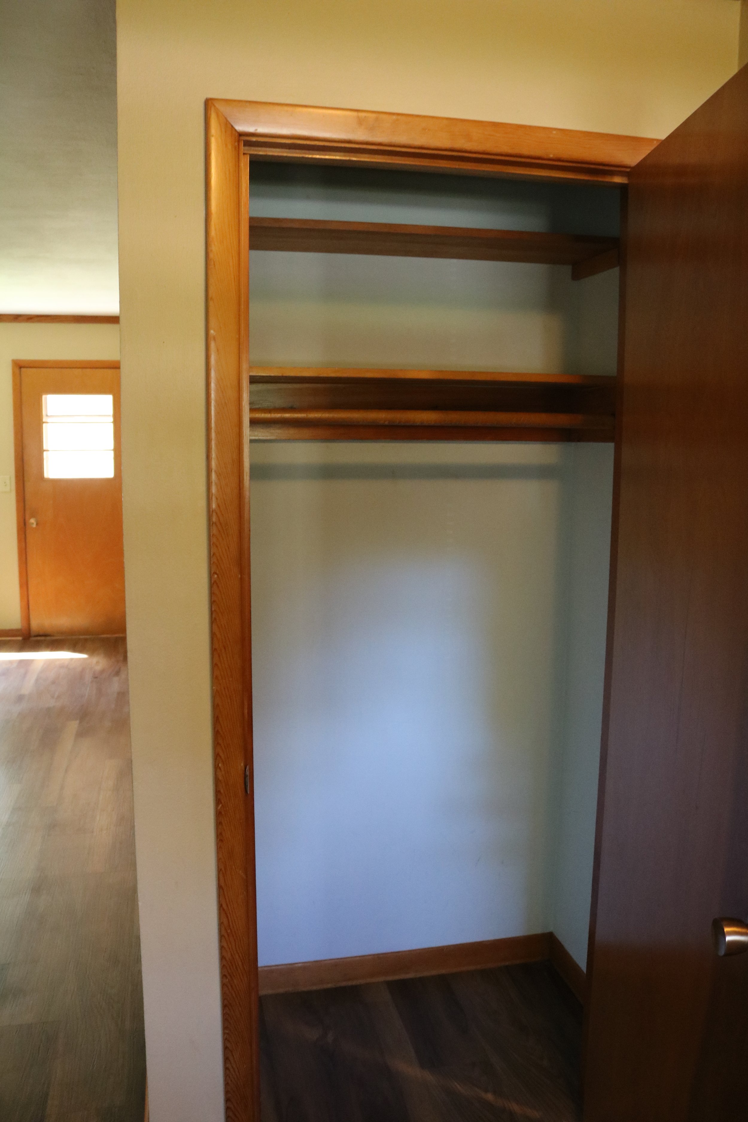 Empty wooden closet with open door, featuring two shelves and a hanging rod, in a room with wooden flooring and a closed door in the background.