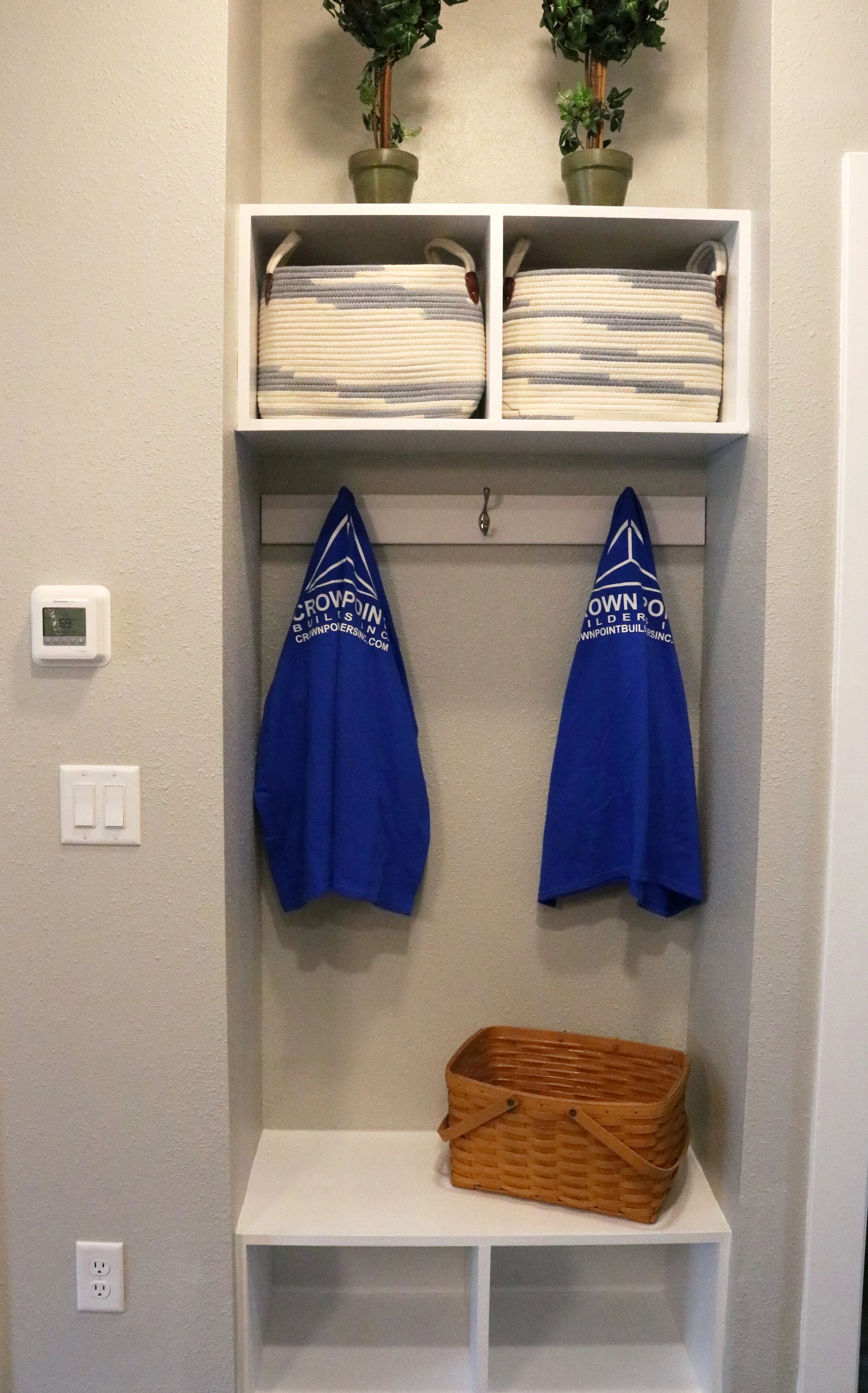 A hallway cubby with two blue jackets hanging on hooks, wicker baskets on shelves above, and a rectangular wicker basket on the lower shelf. There are potted plants on the top shelf and a thermostat and light switches to the left.