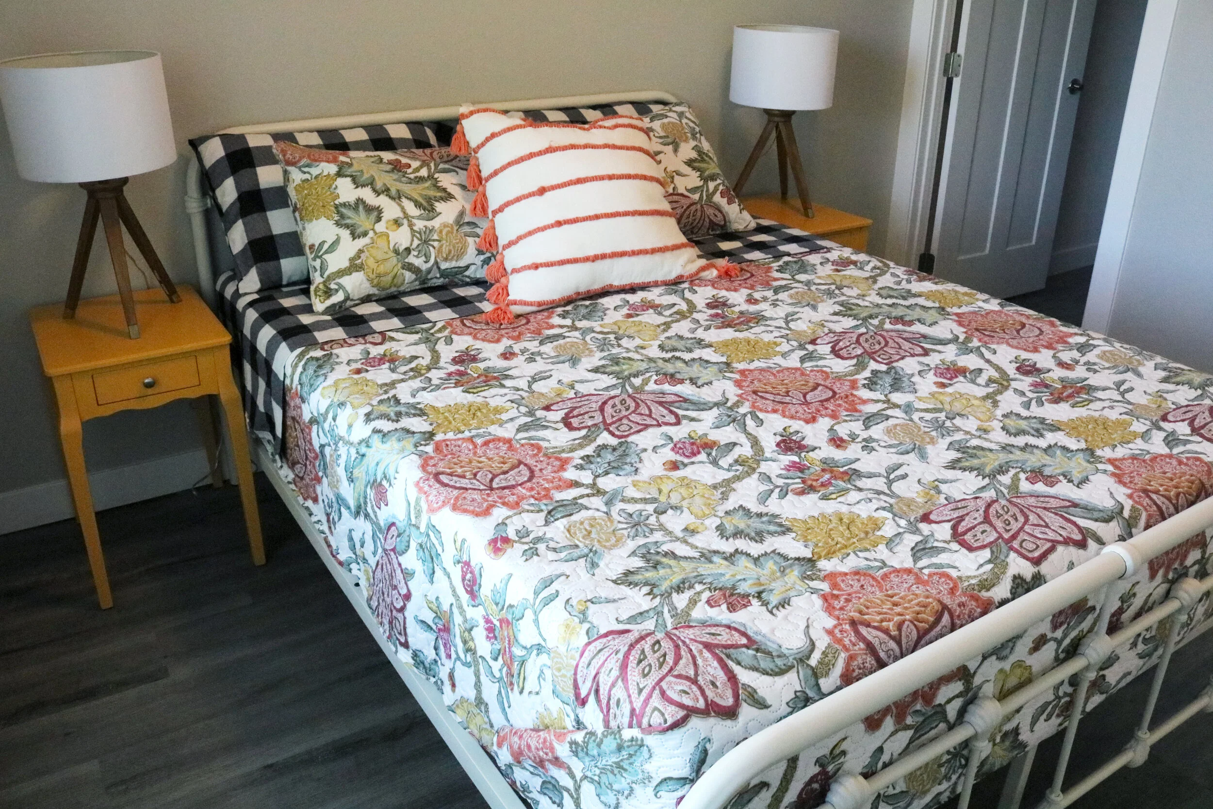 Bedroom with colorful floral-patterned quilt, striped and floral pillows on a bed with black-and-white checked sheets, yellow bedside tables with tripod lamps.