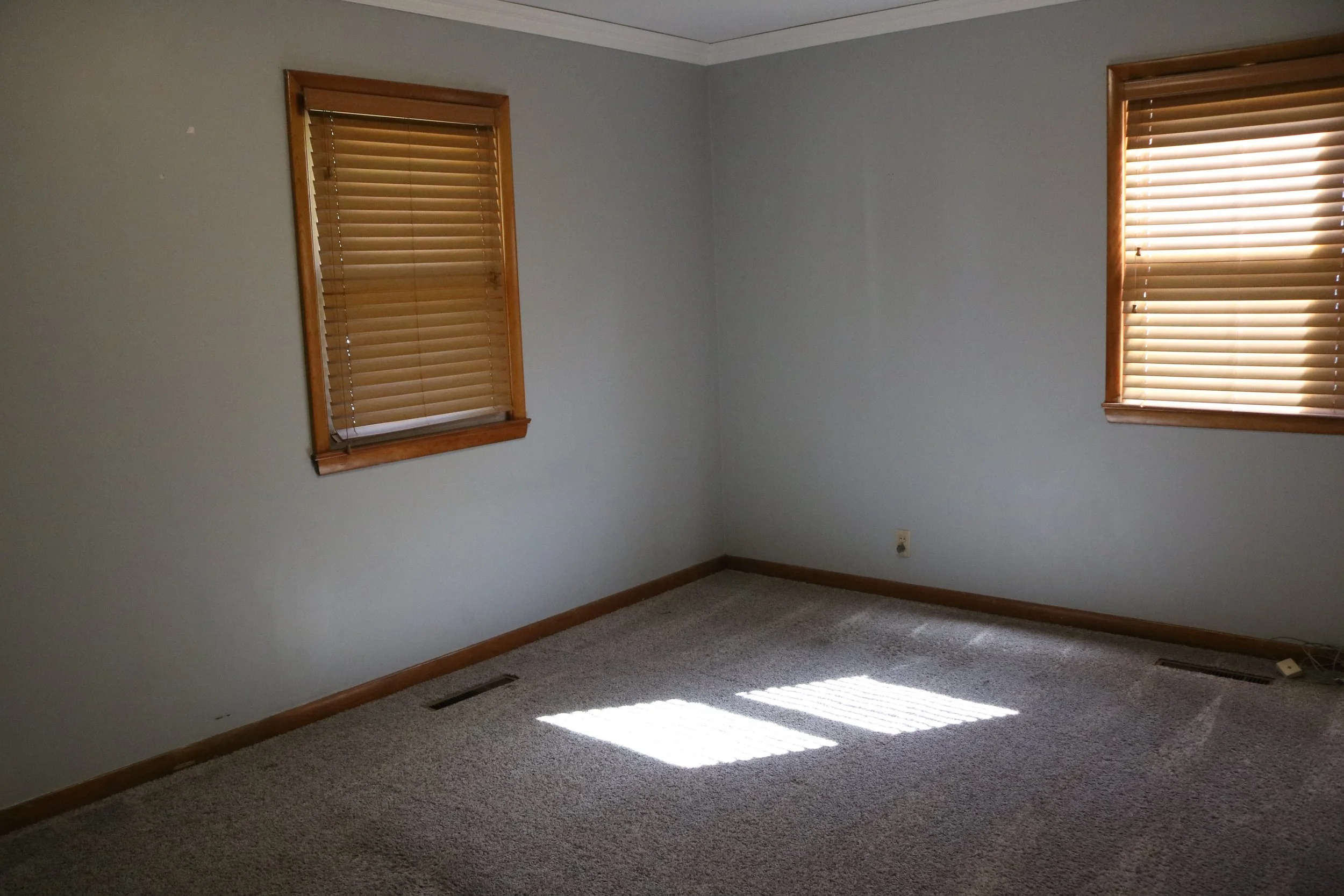 Empty room with light gray walls, two windows with wooden blinds, beige carpet, and sunlight casting shadows on the floor.