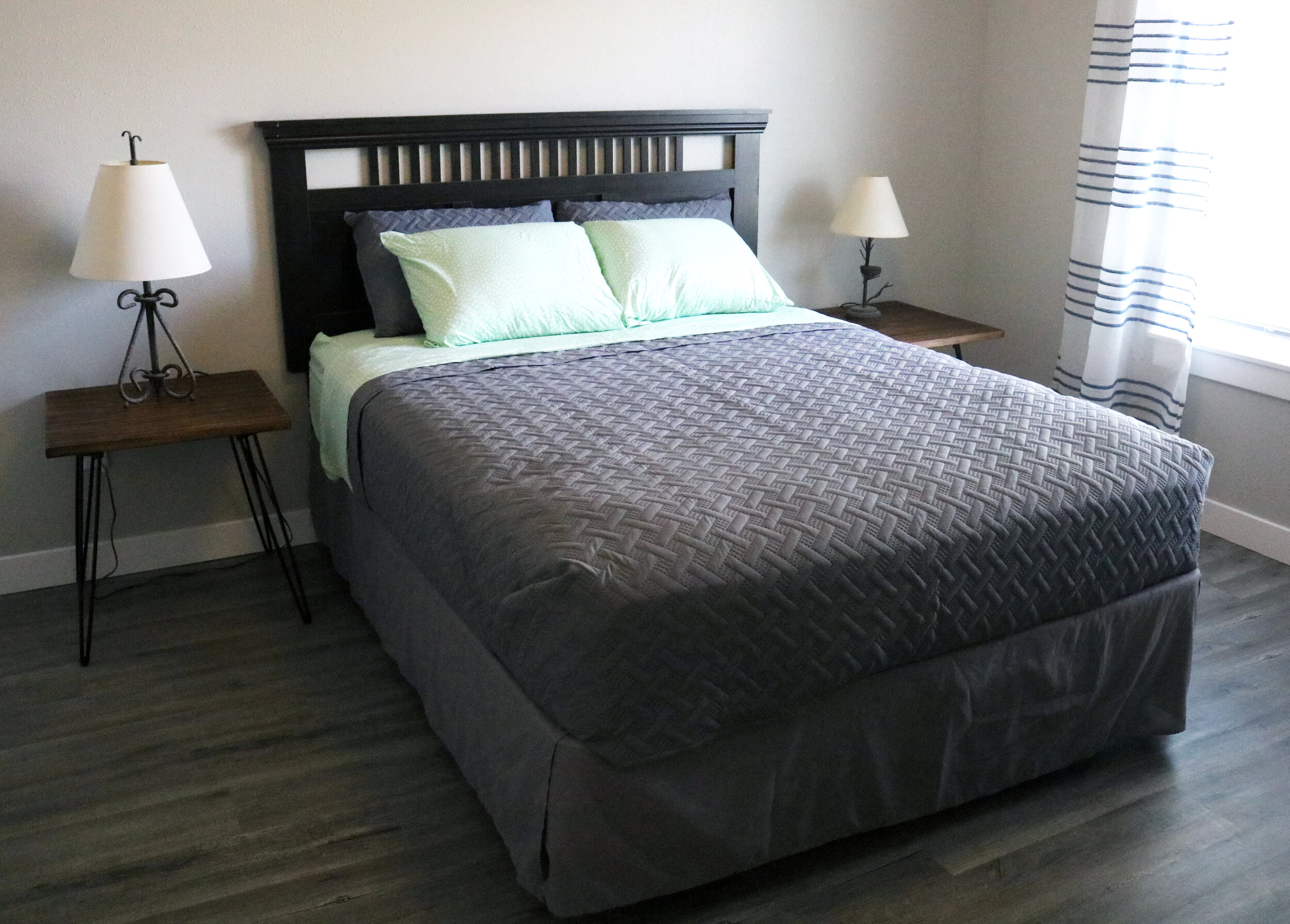 Bedroom with a bed, wooden headboard, gray bedding, two side tables, lamps, and striped curtains.