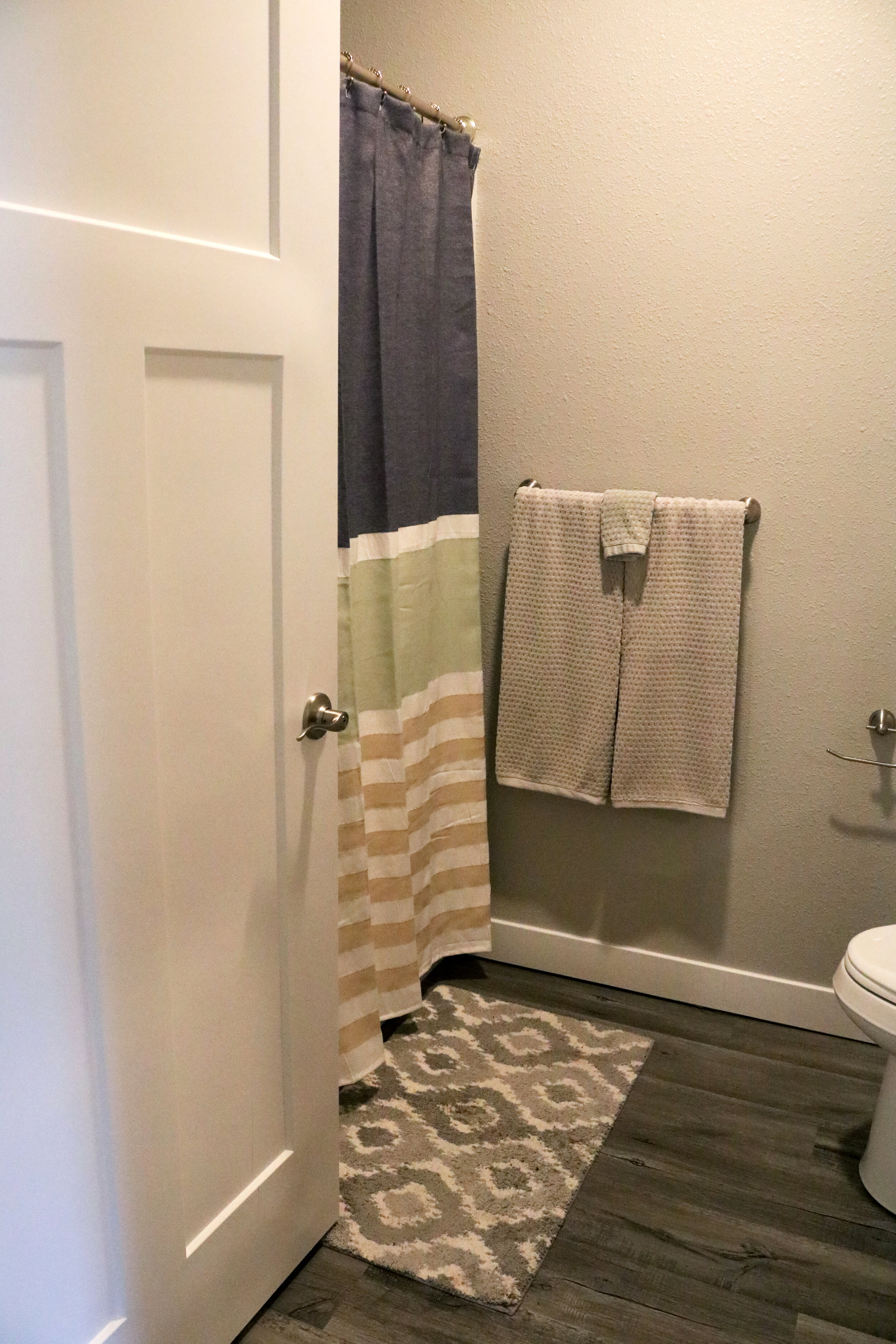 Modern bathroom with a striped shower curtain, patterned rug, gray towels, white door, toilet, and gray walls.