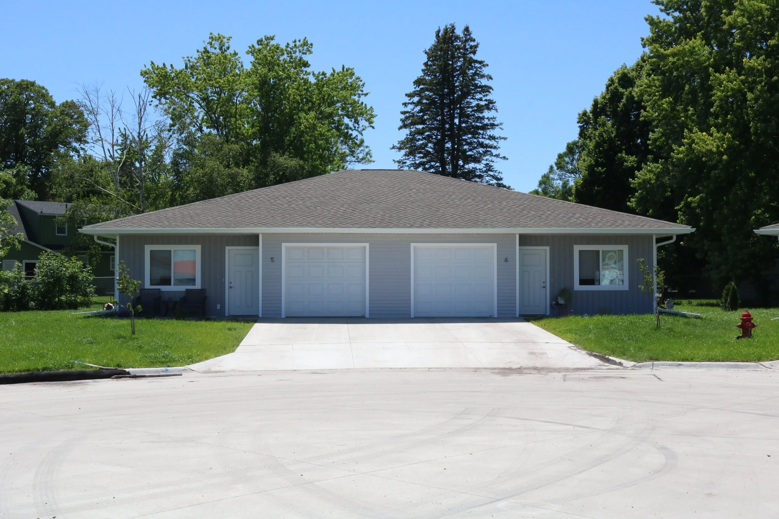 Two-unit duplex with gray siding and two white garage doors, surrounded by green grass and trees.