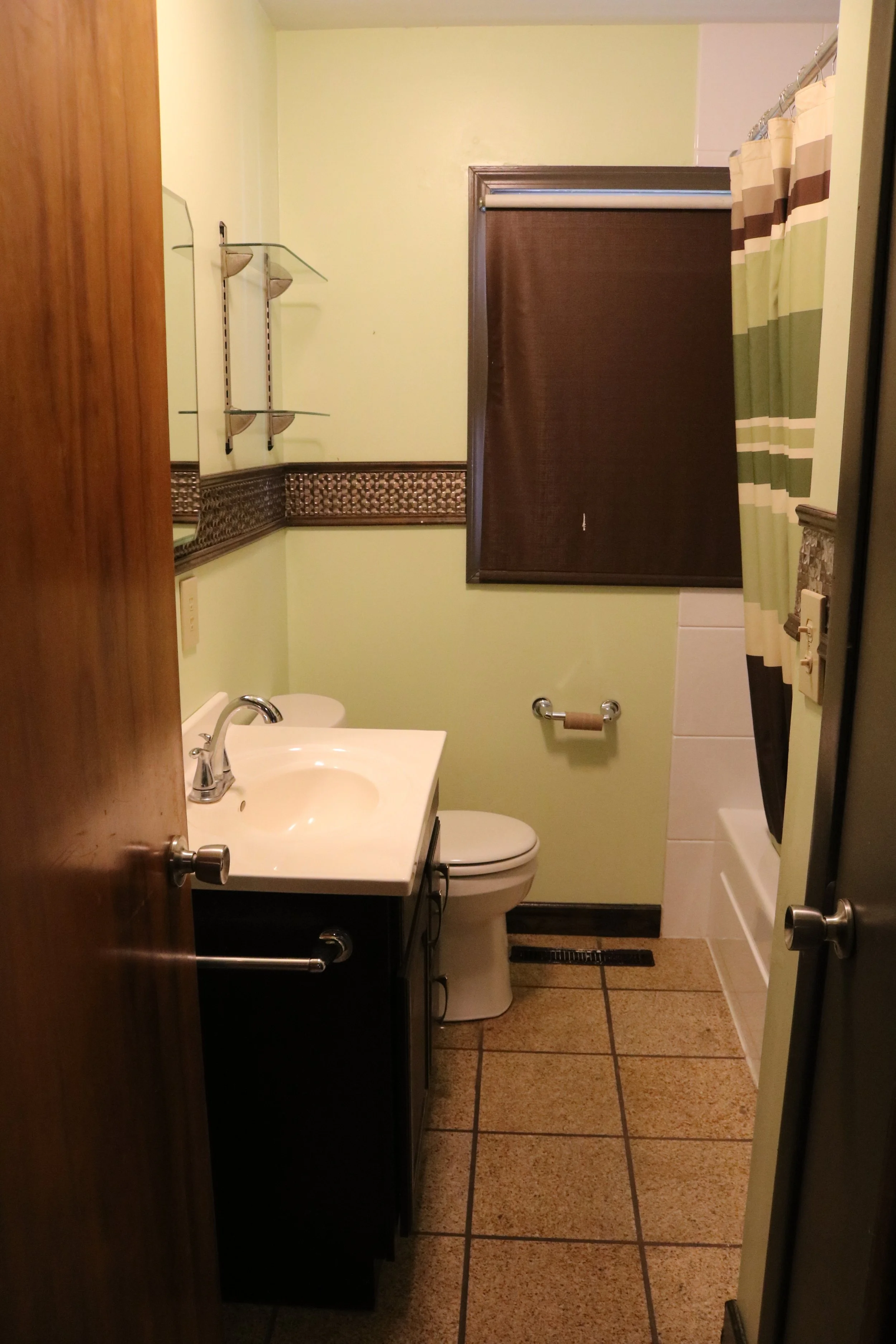 Small bathroom with a vanity sink, toilet, bathtub, and shower curtain. Brown window shade and green walls with decorative tile border. Tiled floor and glass shelves on the wall.