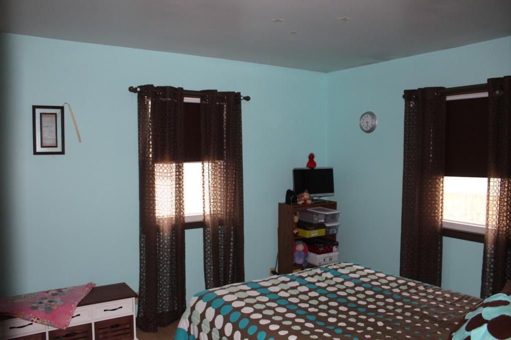 Bedroom with light blue walls, brown polka dot bedspread, brown curtains, and a small TV on a shelf.
