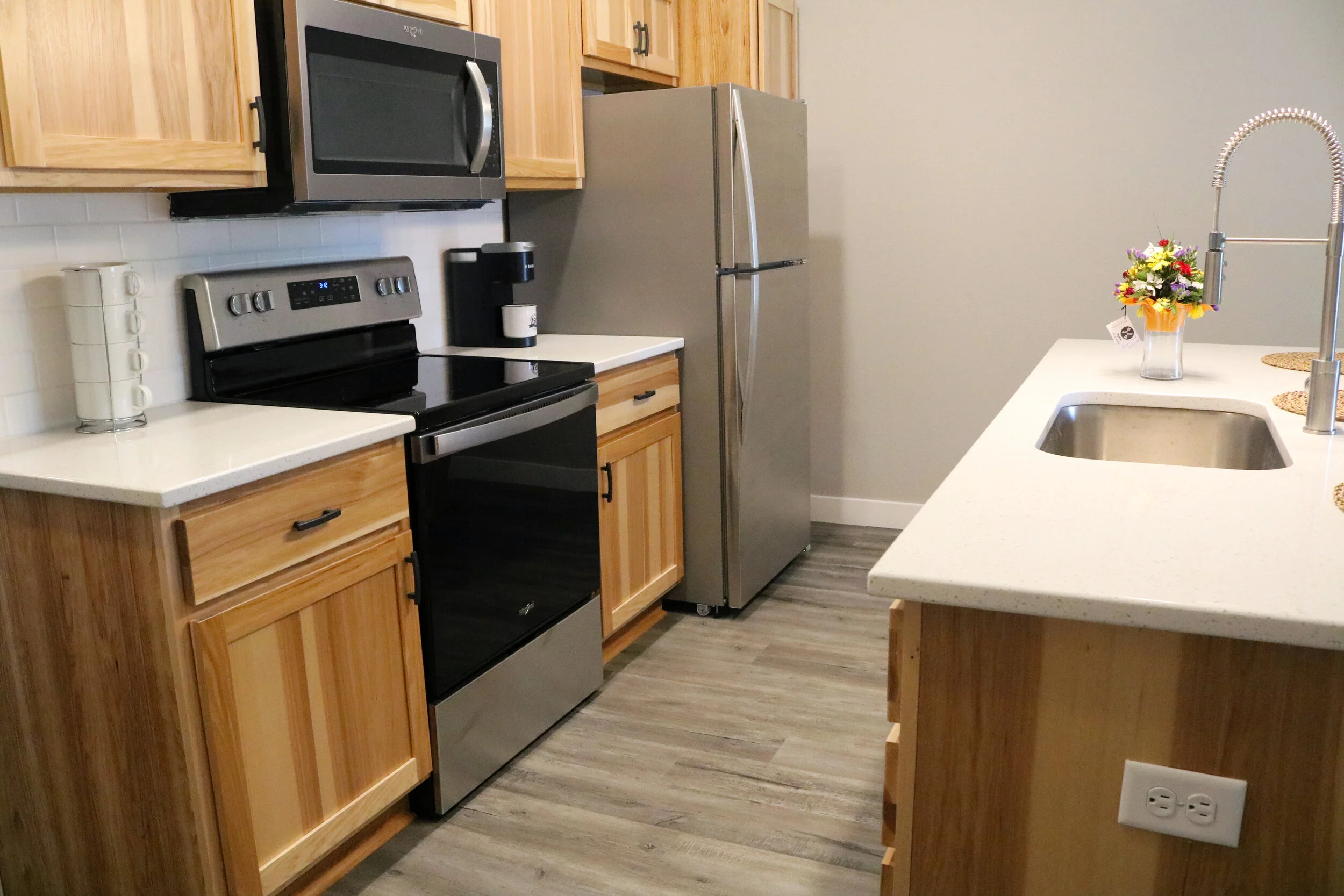 Modern kitchen with stainless steel appliances, light wooden cabinetry, and a central island featuring a sink and decorative vase. The stove and microwave are integrated, with a coffee maker on the countertop. The floor is wooden.