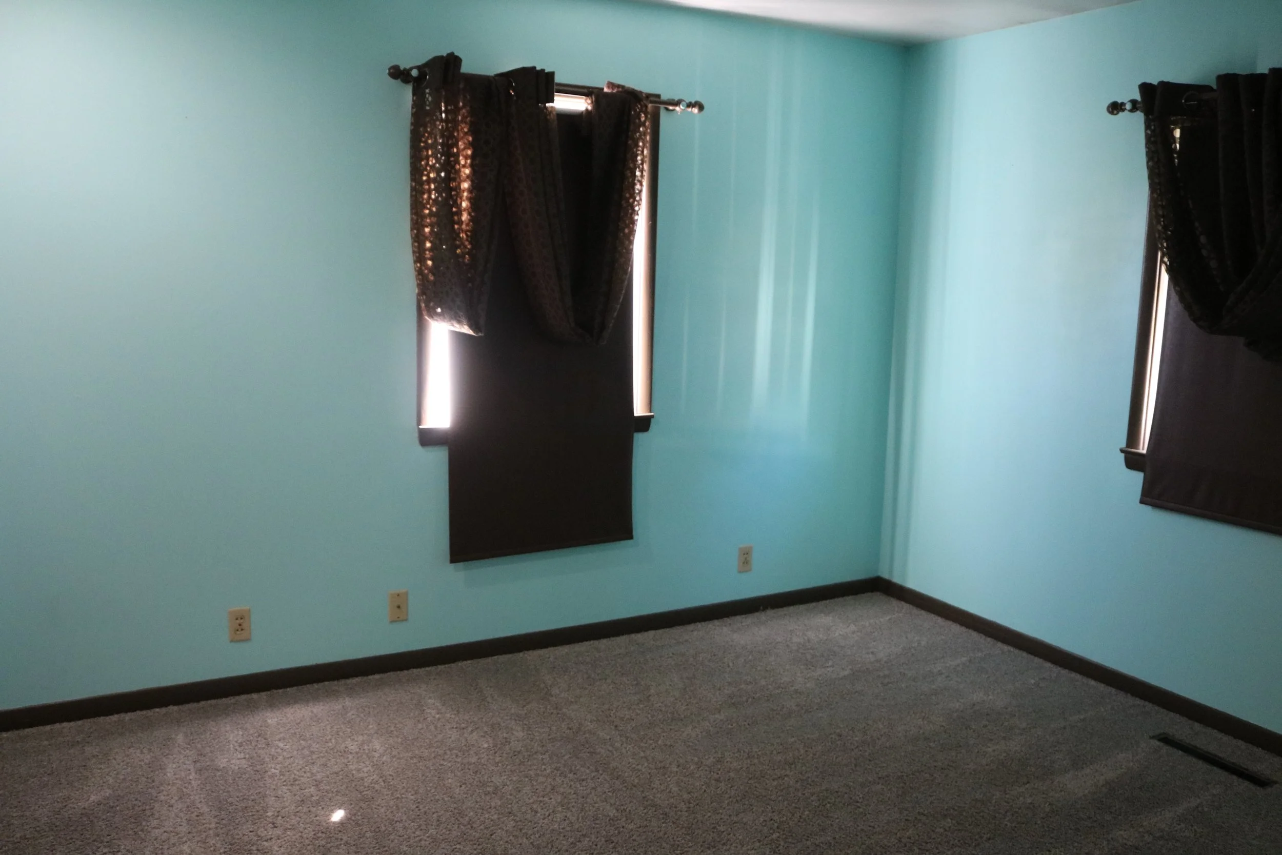 Empty room with turquoise walls, gray carpet, and dark curtains on windows.