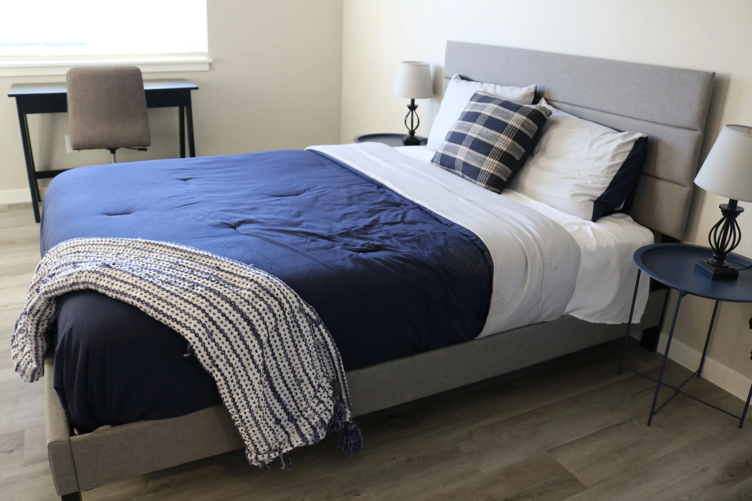 Modern bedroom with gray bed frame, navy bedding, plaid pillows, two black side tables with lamps, and a desk with a chair next to a window.
