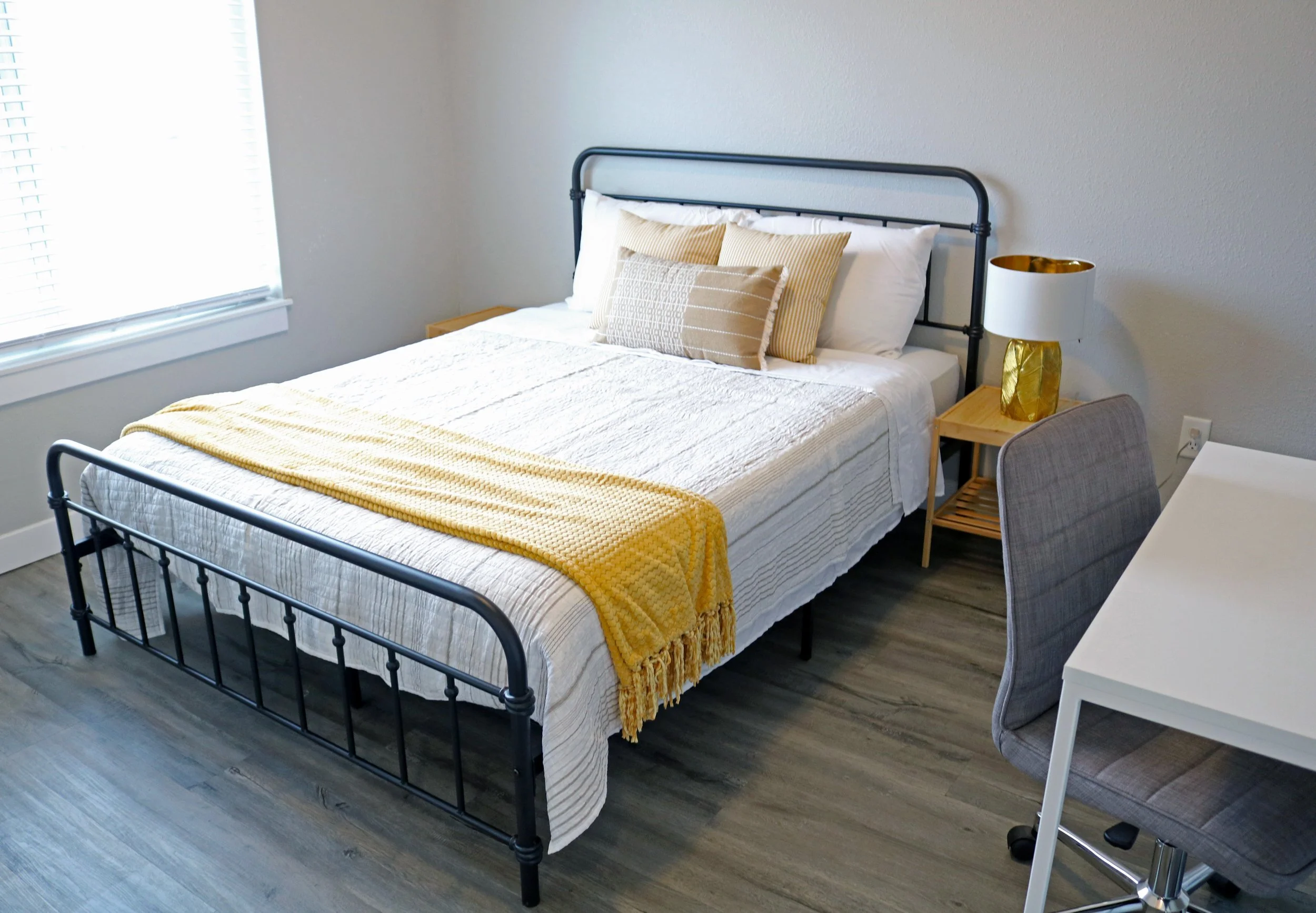 Modern bedroom with a metal bed frame, white bedding and yellow throw blanket, pillows, wooden nightstand with gold lamp, desk and gray chair, and wooden floor.