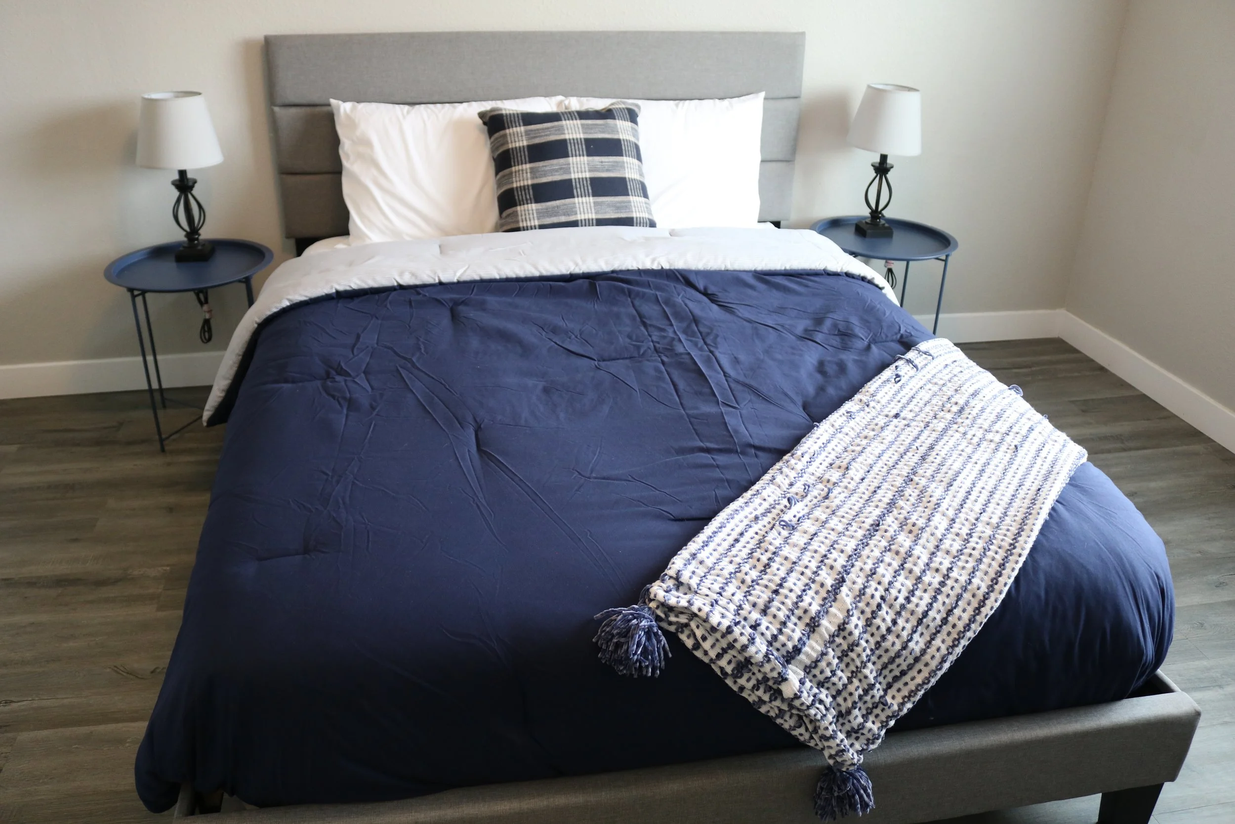 Bedroom with a neatly made bed featuring a navy blue comforter and a blue and white striped throw, flanked by two side tables with lamps.
