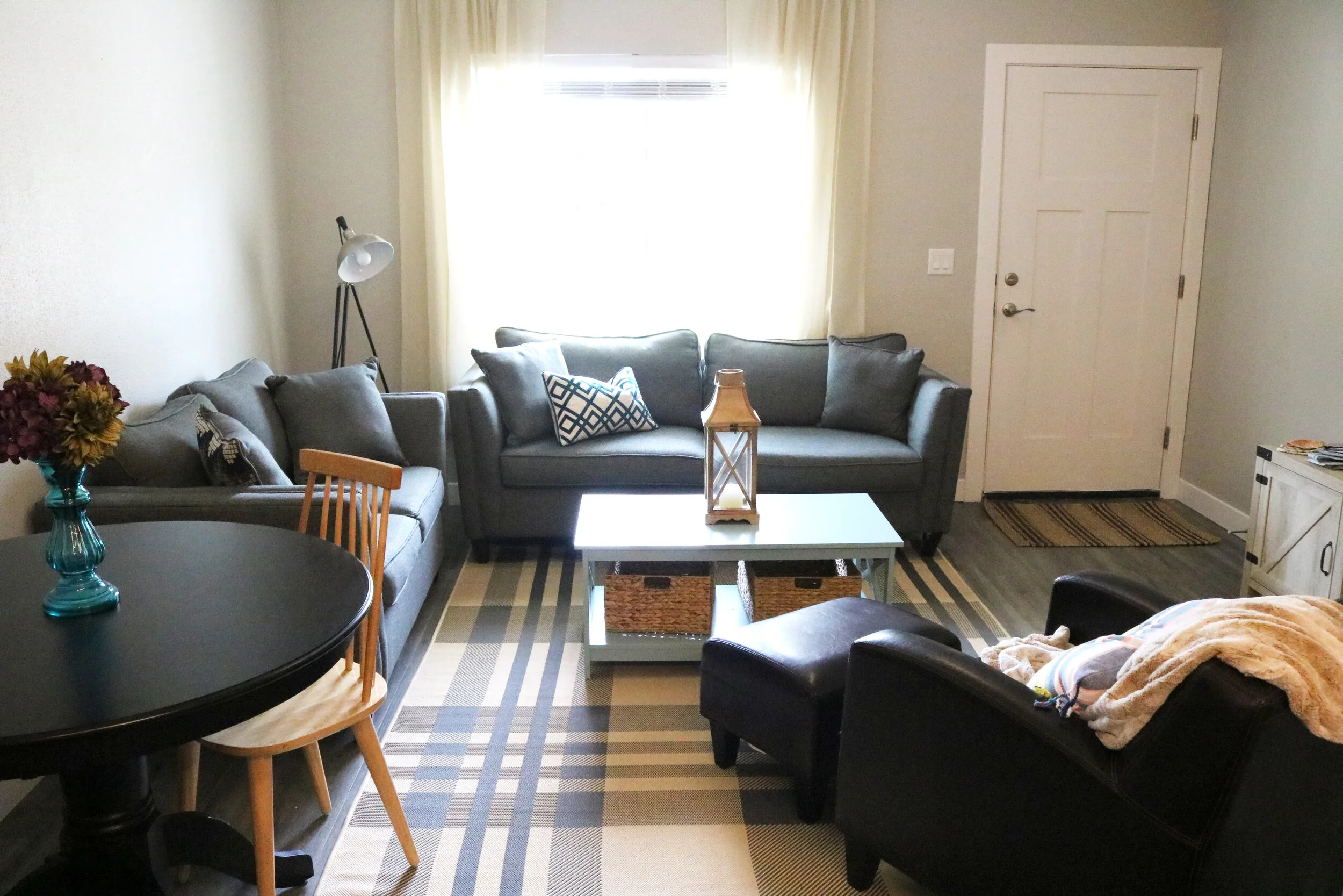 Modern living room with gray sofas, a black round table with a chair, a striped rug, and a blue coffee table with storage baskets. A floor lamp, decorative pillows, and a blanket on a leather chair are present. Neutral color scheme with natural light