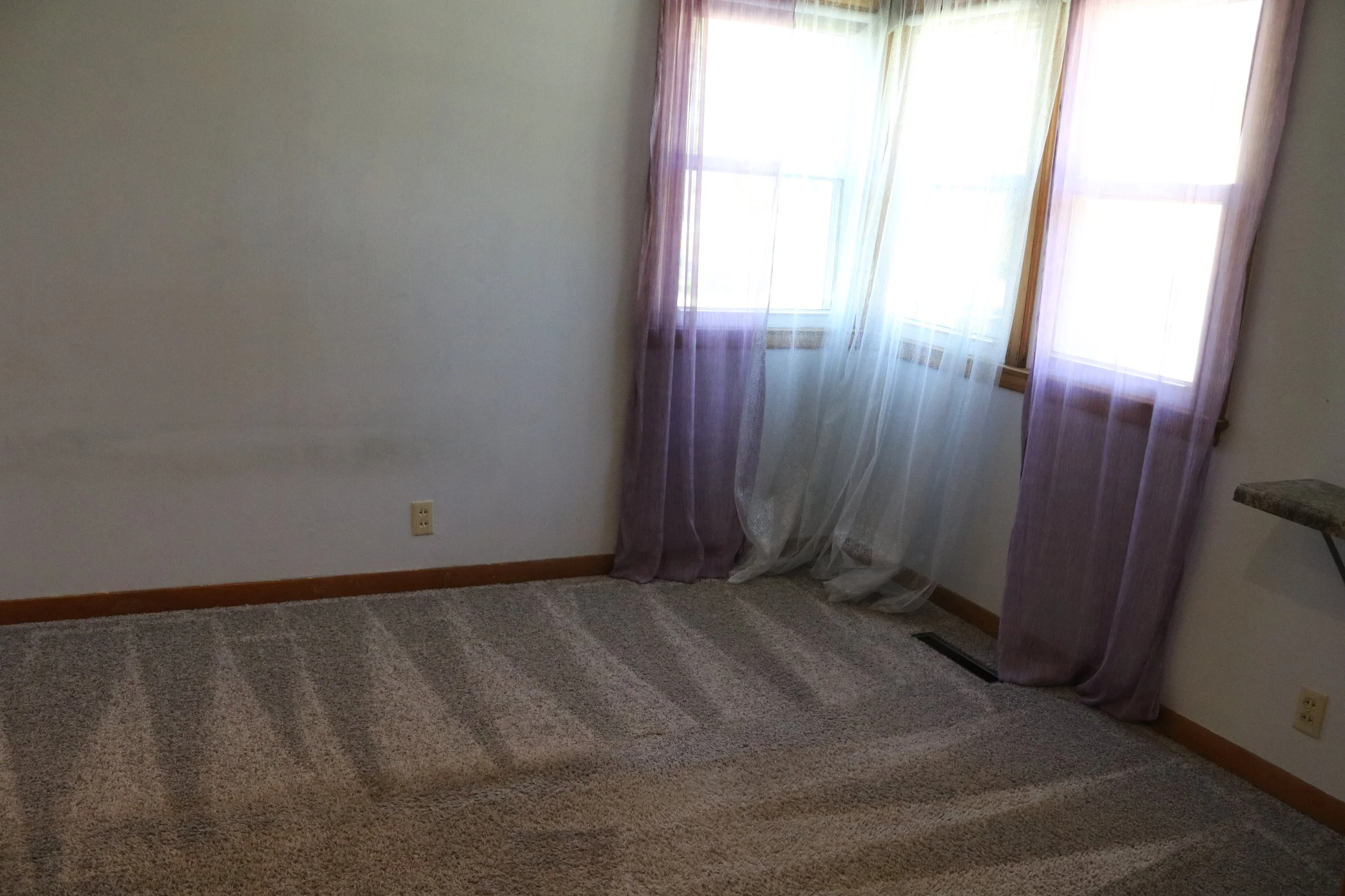 Empty room with beige carpet, two windows with purple sheer curtains, and a heater vent on the floor.