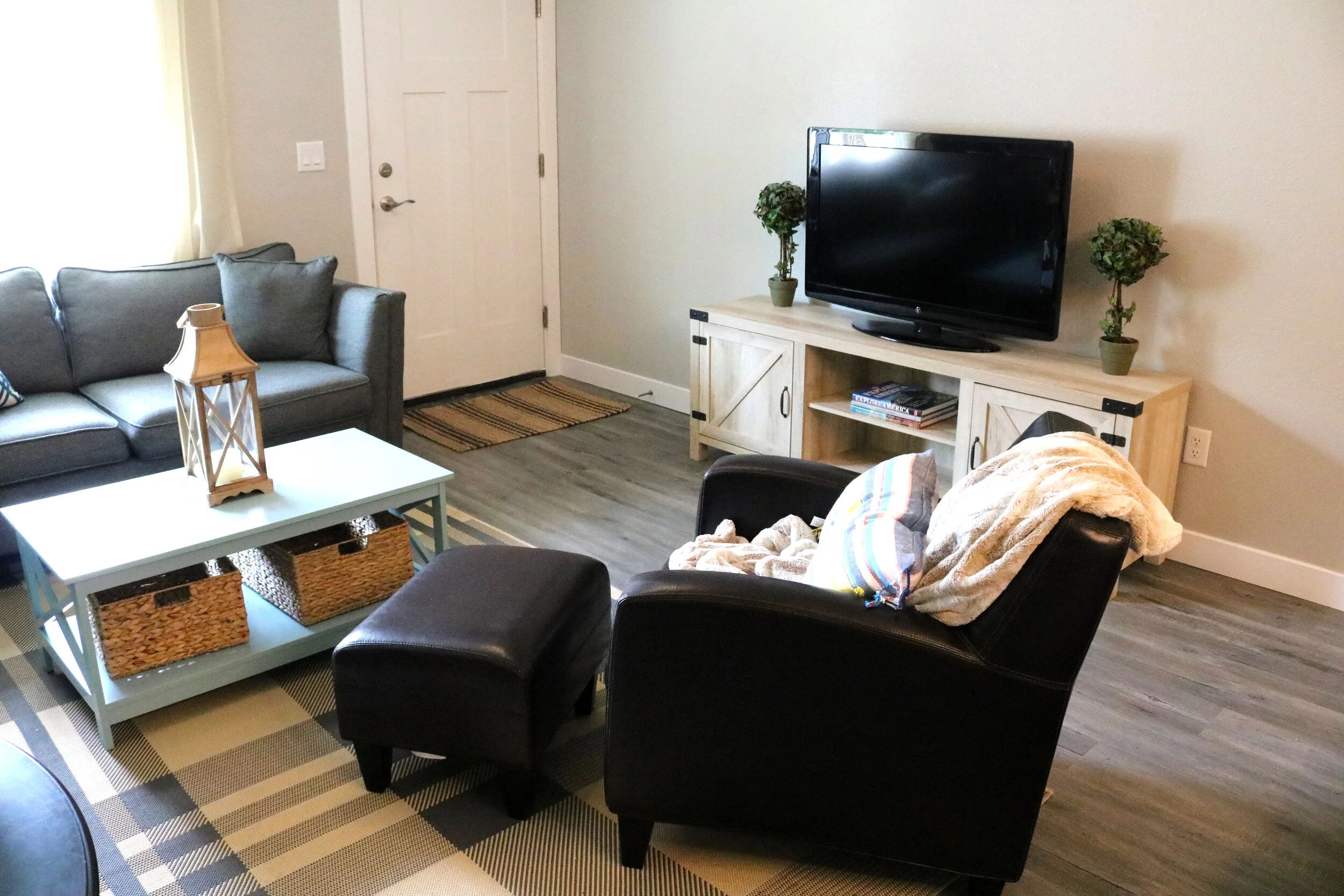 Cozy living room with a gray couch, soft chair, small ottoman, and a TV on a wooden stand. Light blue coffee table with woven baskets underneath and lantern decor on top. Neutral-colored rug.