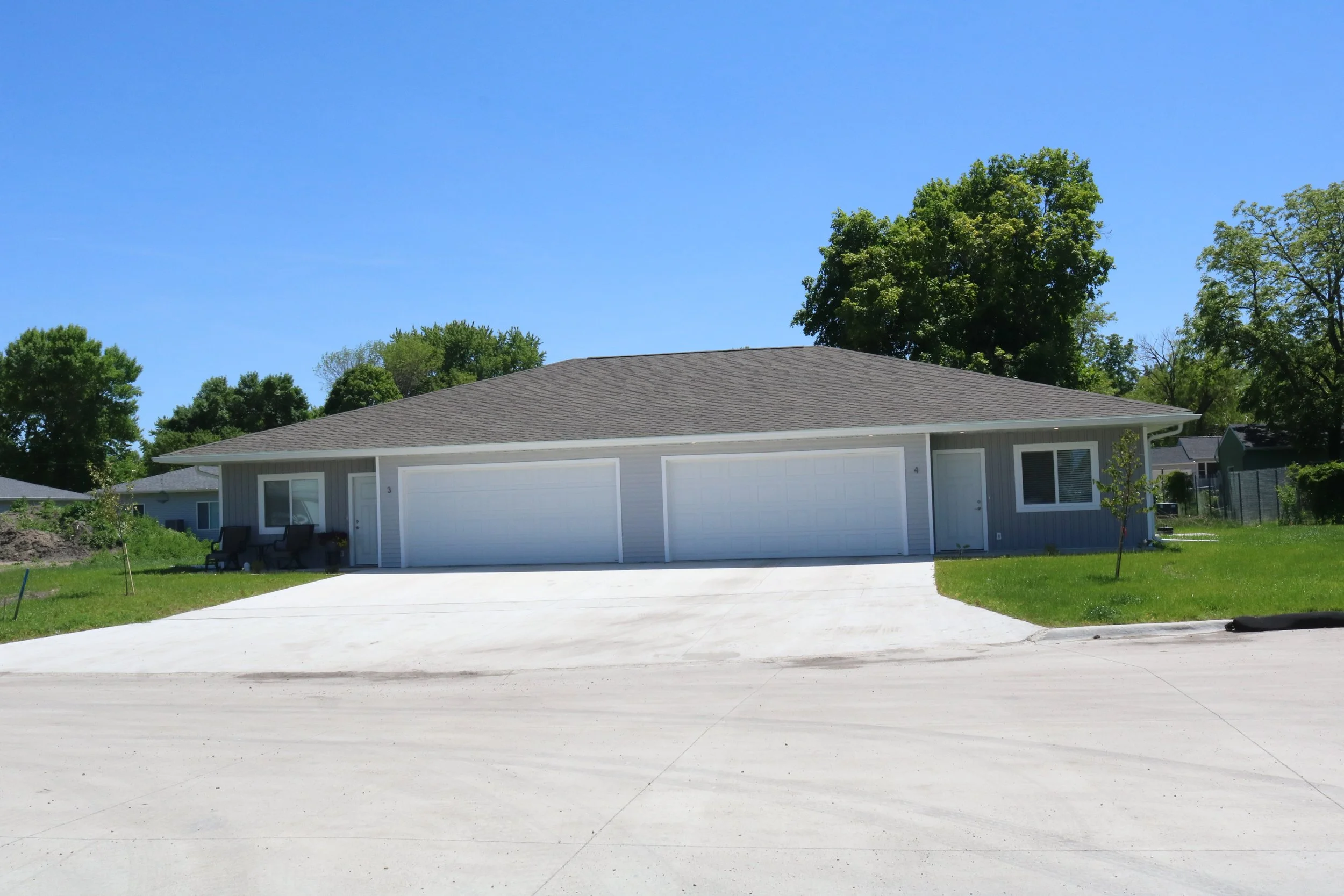 A single-story duplex with two white garage doors in front, gray siding, a paved driveway, and a grassy lawn with trees in the background under a clear blue sky.