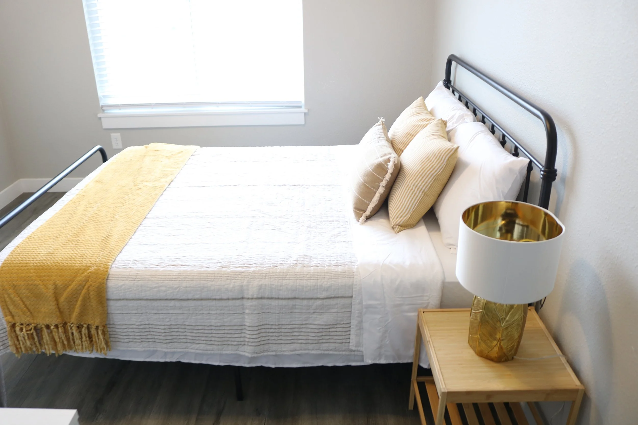 Bedroom with a neatly made bed featuring white sheets and two decorative pillows, a yellow throw blanket, a black metal bed frame, and a wooden nightstand holding a gold and white table lamp.