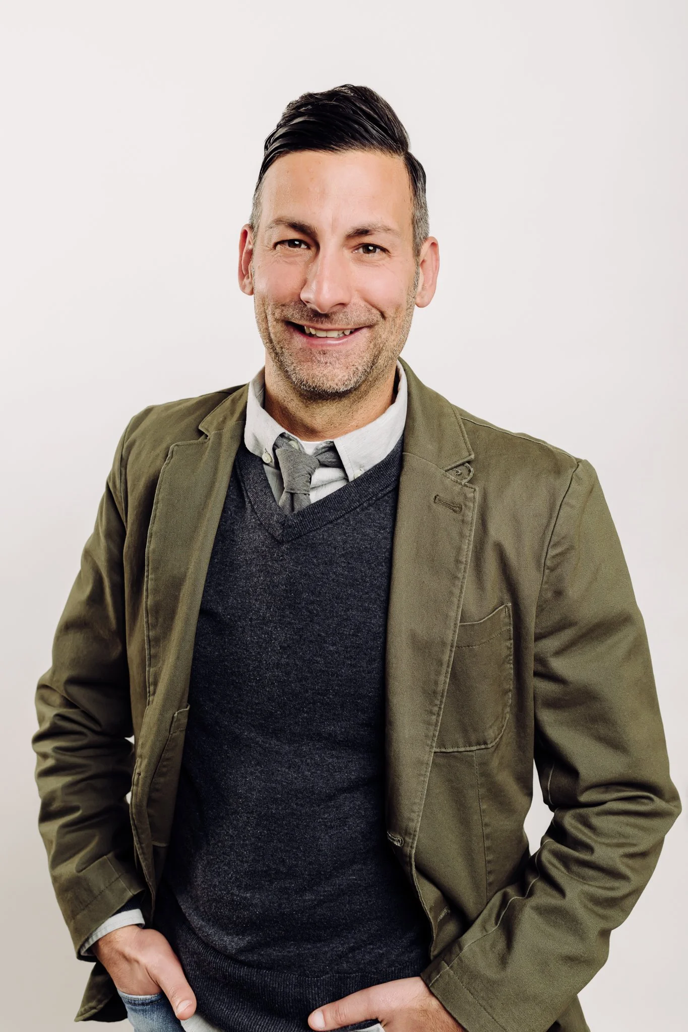 A man with short dark hair and stubble, wearing a grey shirt, grey sweater, and a khaki jacket, smiling against a plain white background.