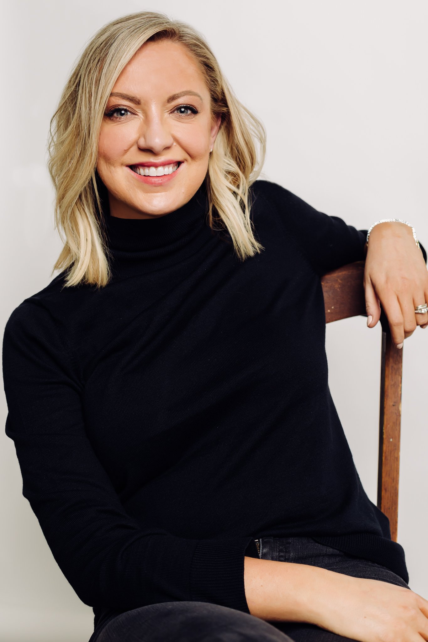 A smiling blonde woman in a black turtleneck sitting on a wooden chair with a plain white background.