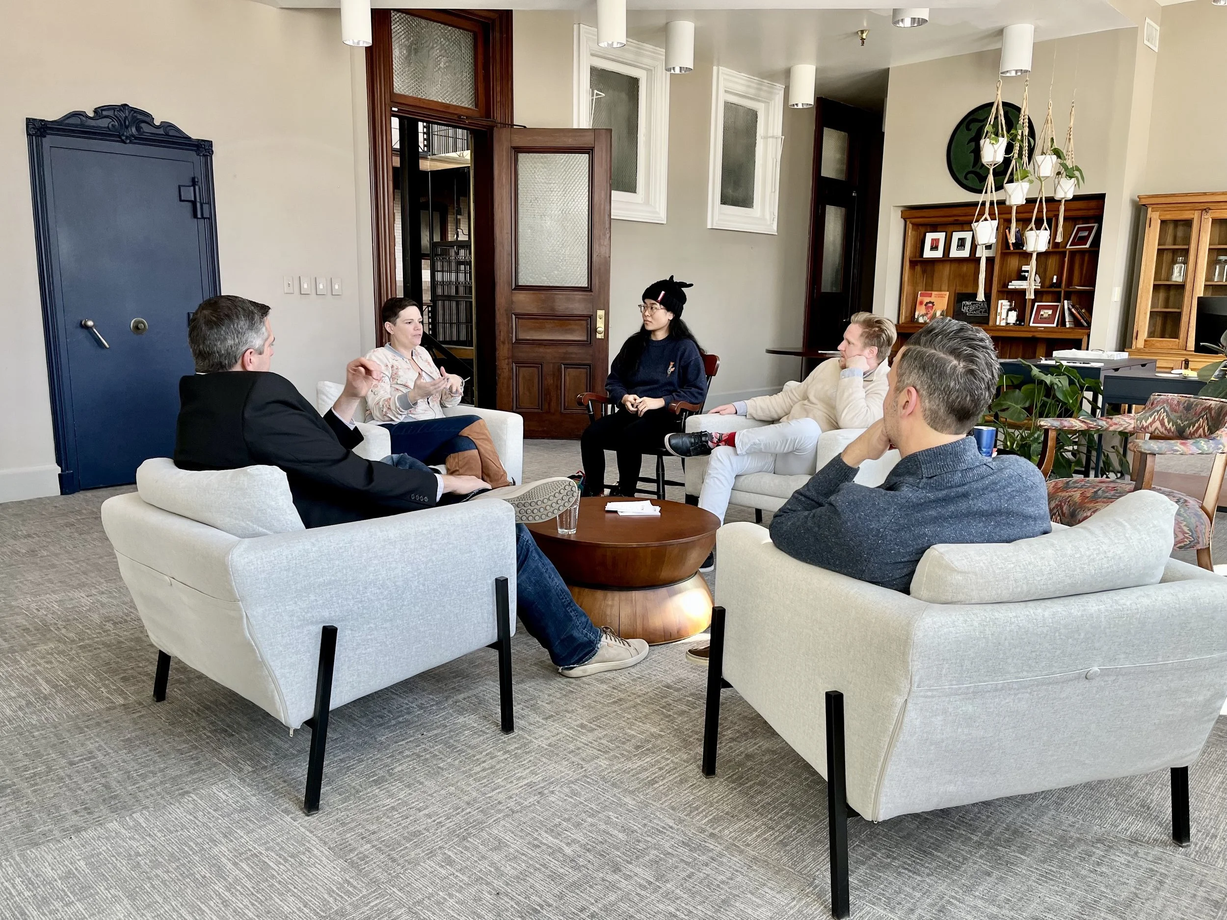 A group of six people having a discussion in a modern living room with white sofas, wooden shelves, and potted plants.