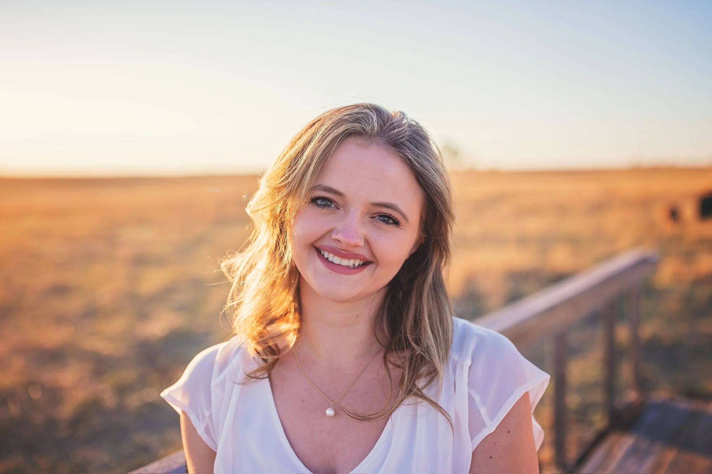 A young woman with light brown hair and blue eyes smiling outdoors at sunset, wearing a white top and a pearl pendant necklace.