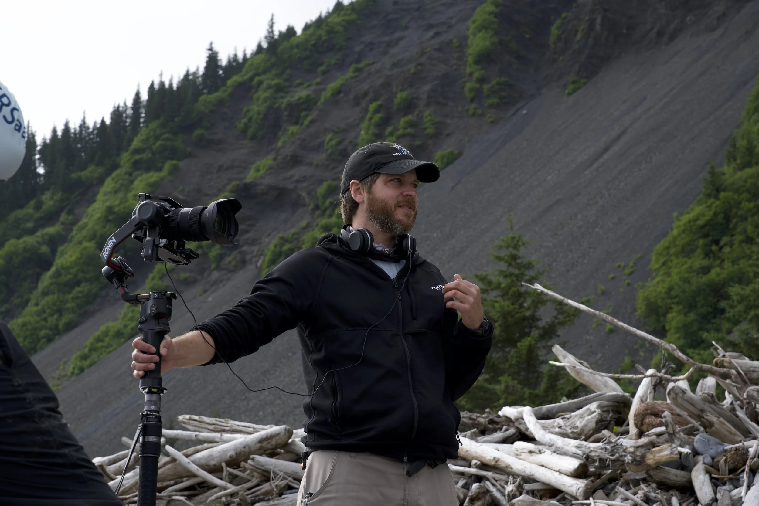A man with a beard wearing a black jacket, baseball cap, and headphones around his neck holding a camera on a stabilizer, outdoors with a forested hillside and driftwood in the background.