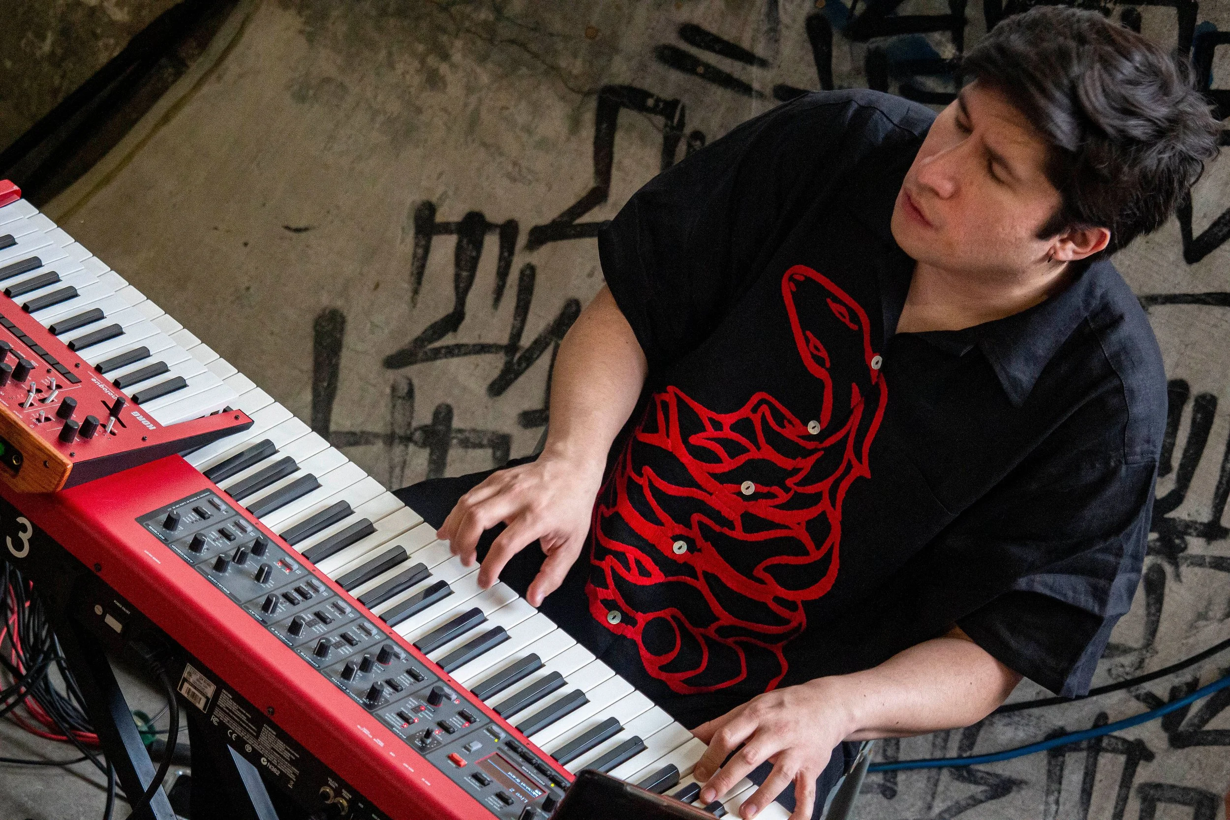 A person playing a red electronic keyboard with various control knobs, wearing a black shirt with a red graphic design, in front of a graffiti-style wall.