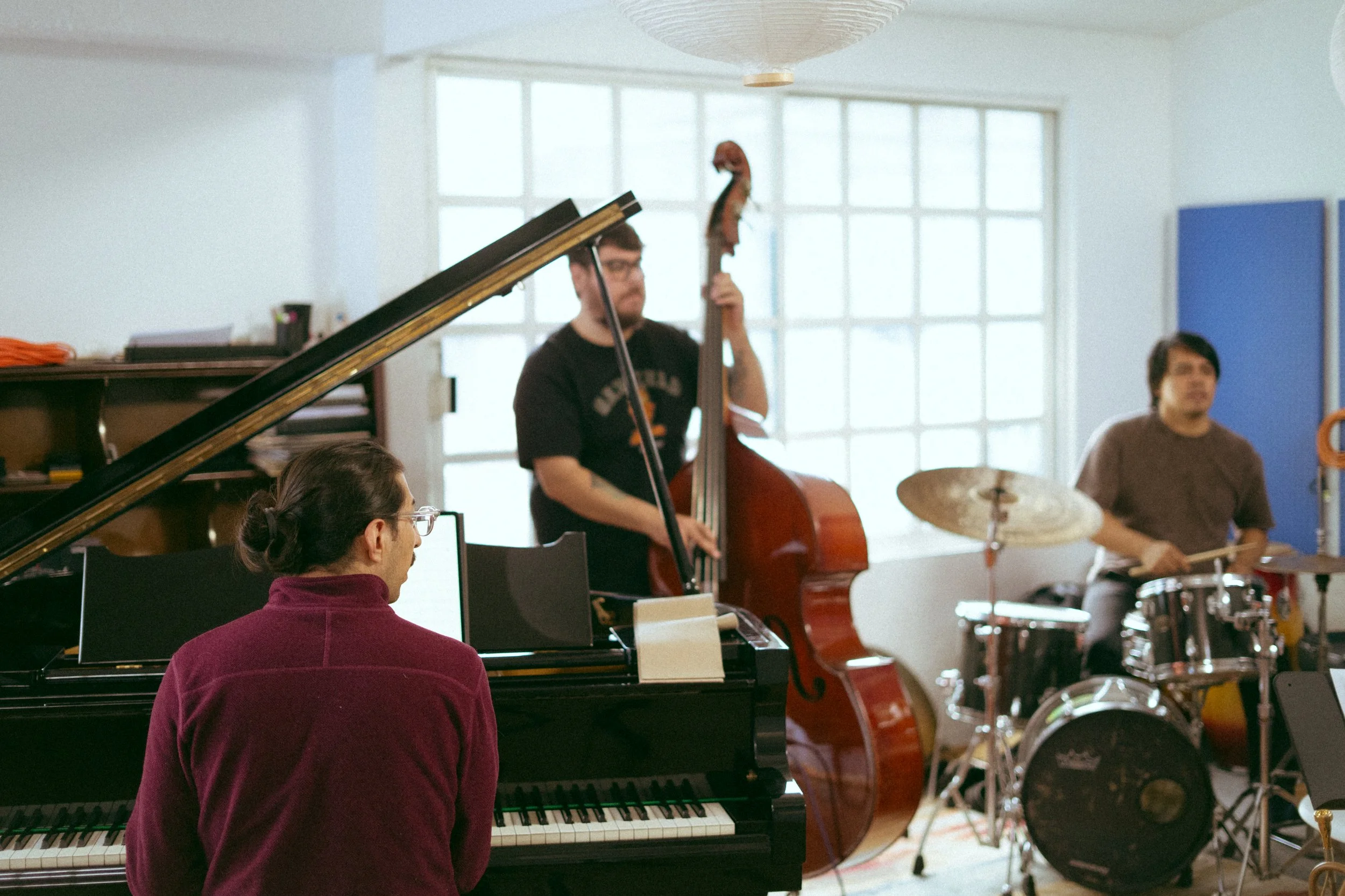Three musicians playing instruments in a brightly lit room: a woman playing the piano, a man playing the double bass, and another man playing the drums.