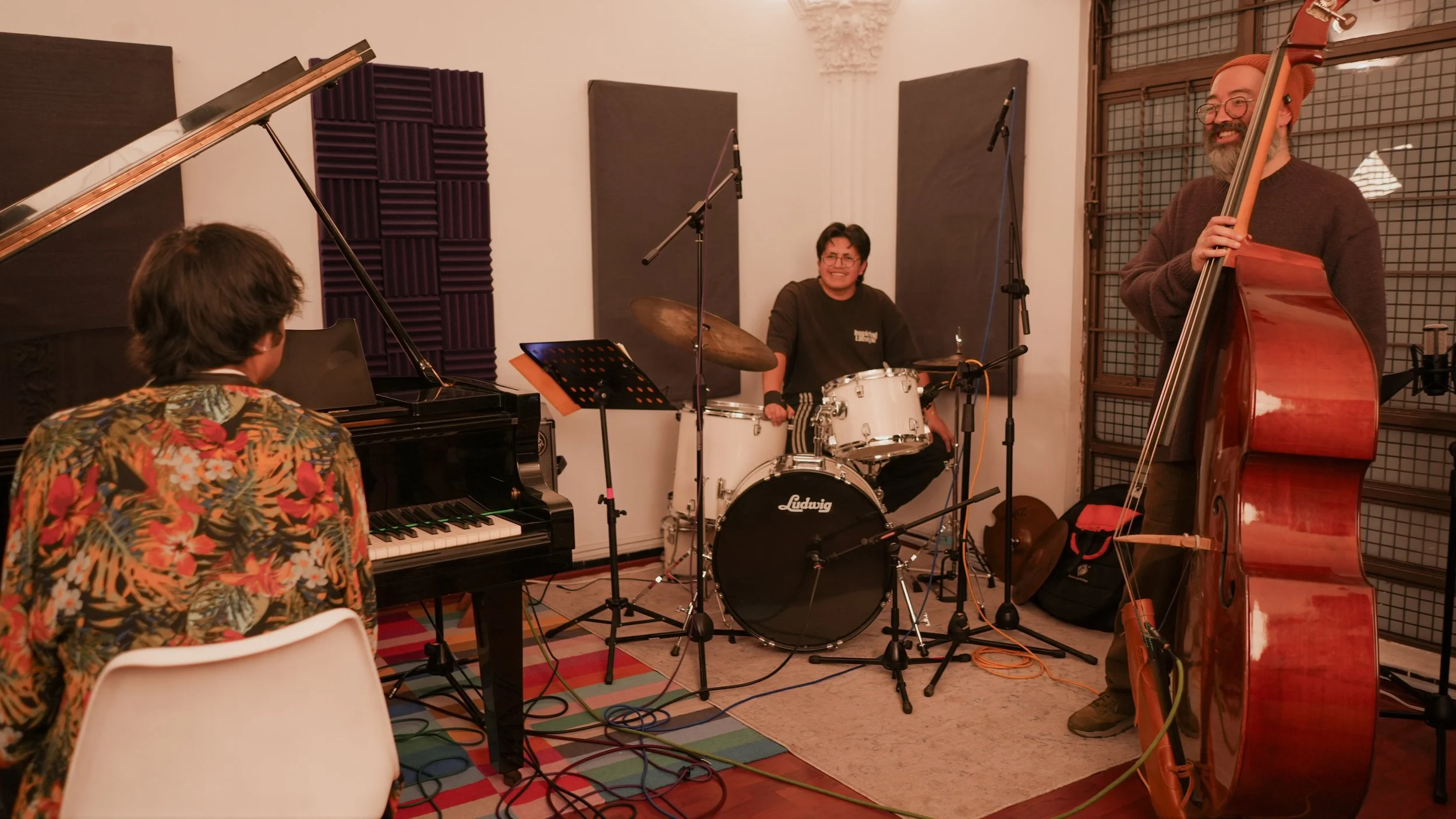 Three musicians playing instruments in a brightly lit room: a woman playing the piano, a man playing the double bass, and another man playing the drums.