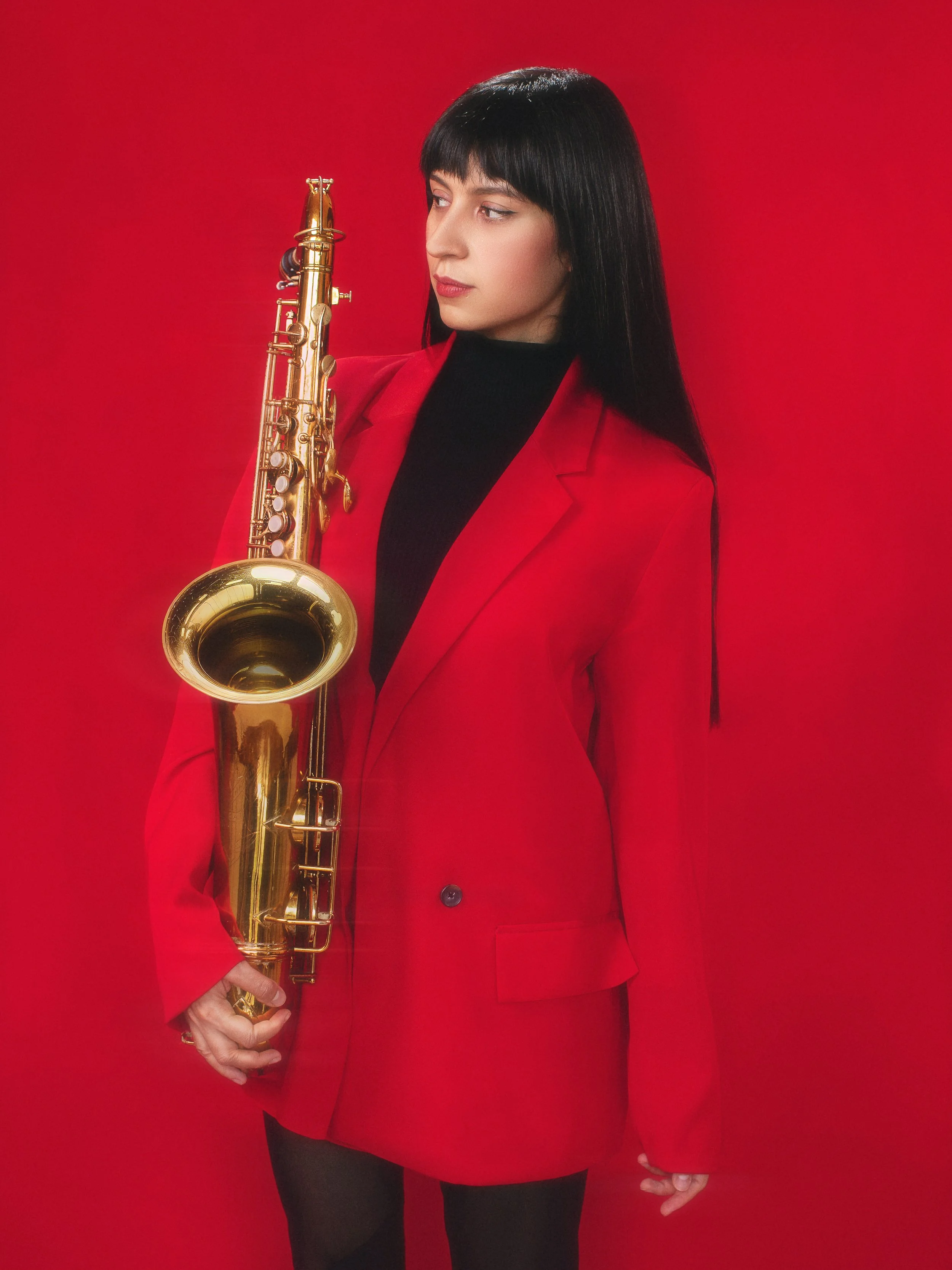 A woman with long black hair wearing a red blazer and black turtleneck holding a gold saxophone against a red background.