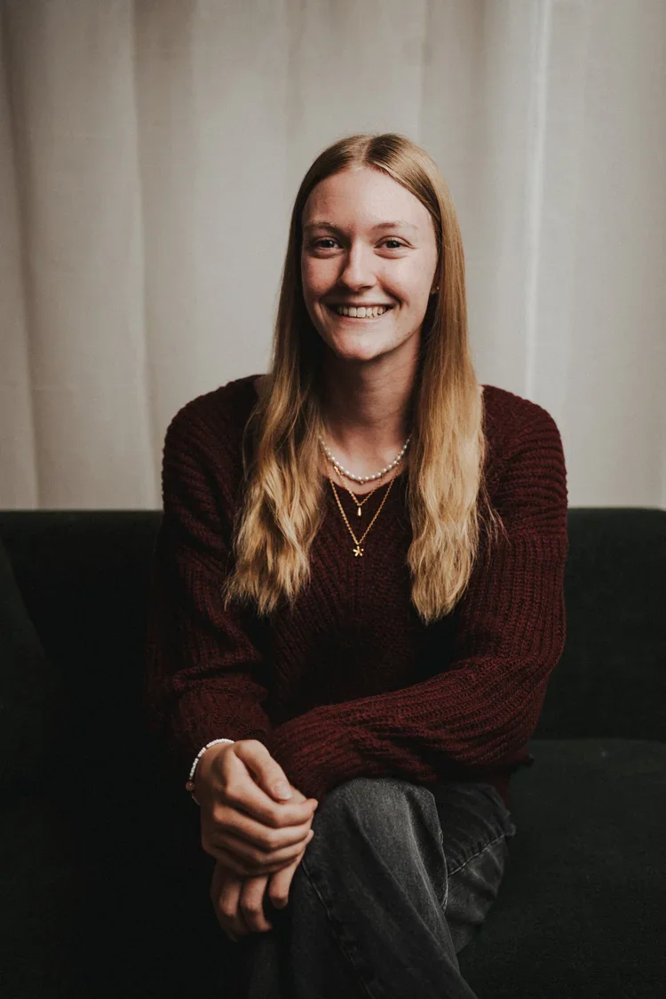 Junge Frau mit langen blonden Haaren, lächelnd, trägt dunkle Kleidung und Schmuck, sitzt auf einem schwarzen Sofa vor einem weißen Vorhang.