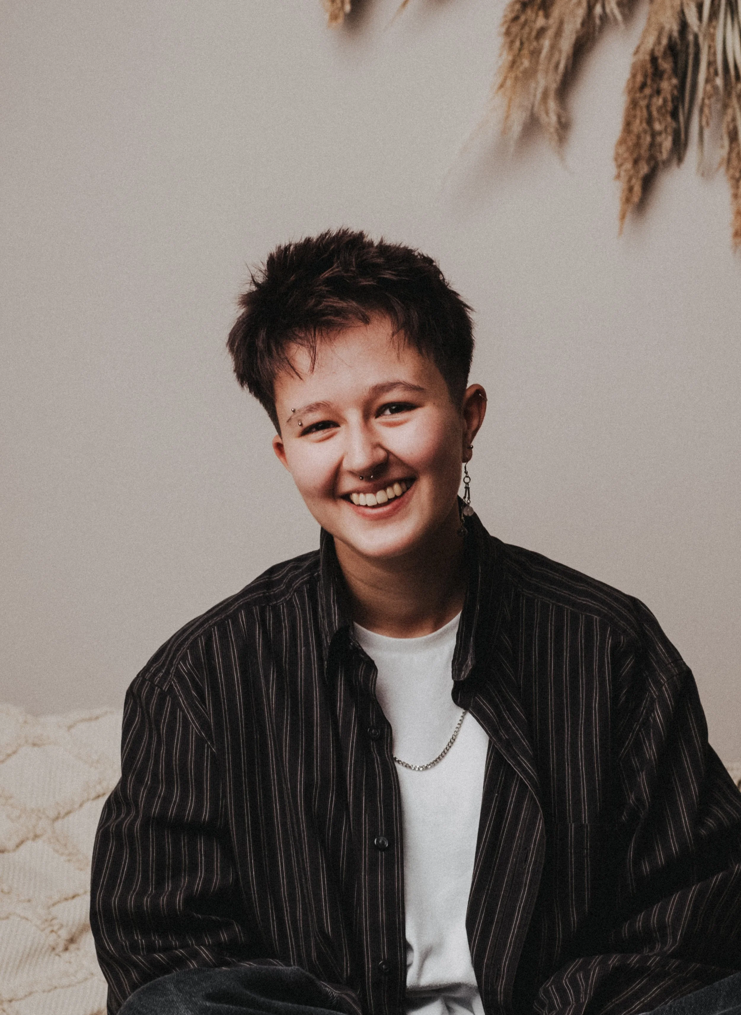 Junge mit kurzen dunklen Haaren, Piercings, Ohrringen und einer Halskette, trägt ein schwarzes gestreiftes Hemd und sitzt auf einem Sofa, lachend.