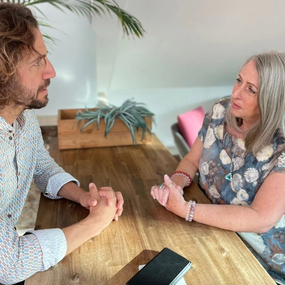 Two people sitting across from each other at a wooden table, engaged in conversation. One person gestures with their hands, and a potted plant is in the background.