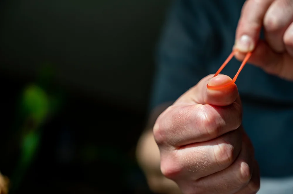 A hand stretching an orange rubber band over a thumb, ready to launch it