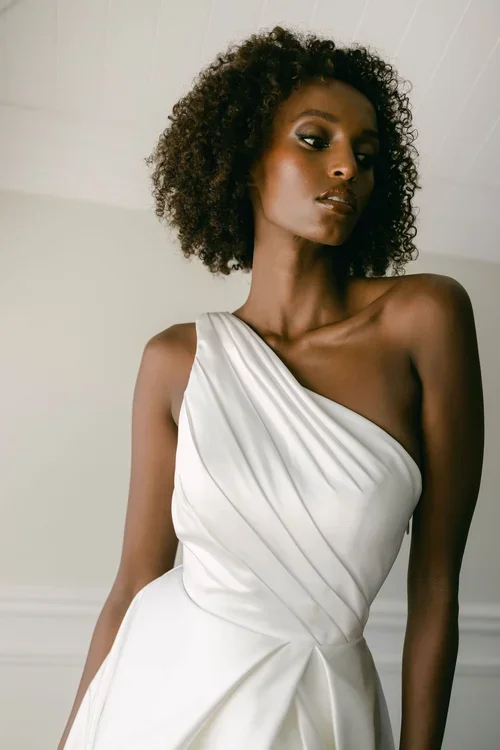 A woman with curly hair wears one-shoulder, EB22531-1Z white pleated dress, standing indoors against a light-colored wall and looking slightly away from the camera.