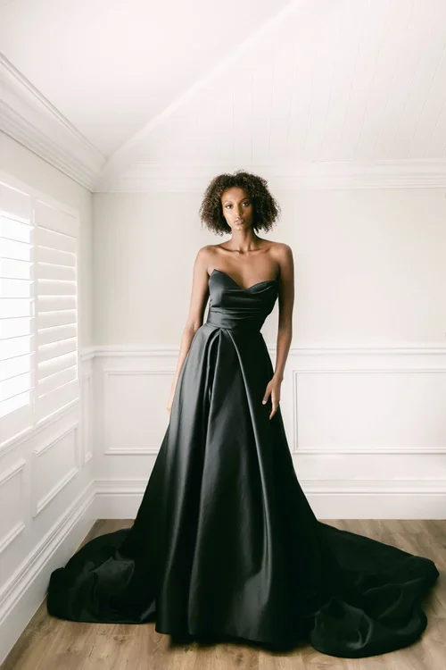 A woman stands in a bright, white room wearing EB22456-1Z strapless, floor-length black gown with a full skirt and a slight train, her hair styled in natural curls.
