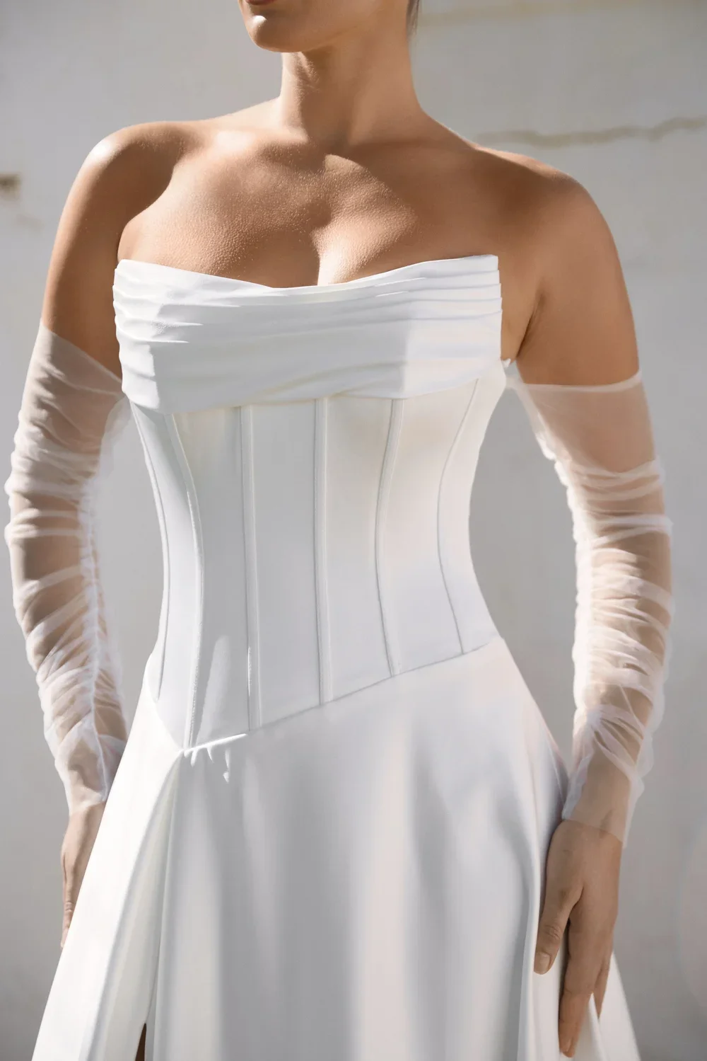 A woman wearing an elegant white off-the-shoulder Everlyn dress with a fitted, corset-style bodice and sheer long sleeves, standing against a light, neutral background.