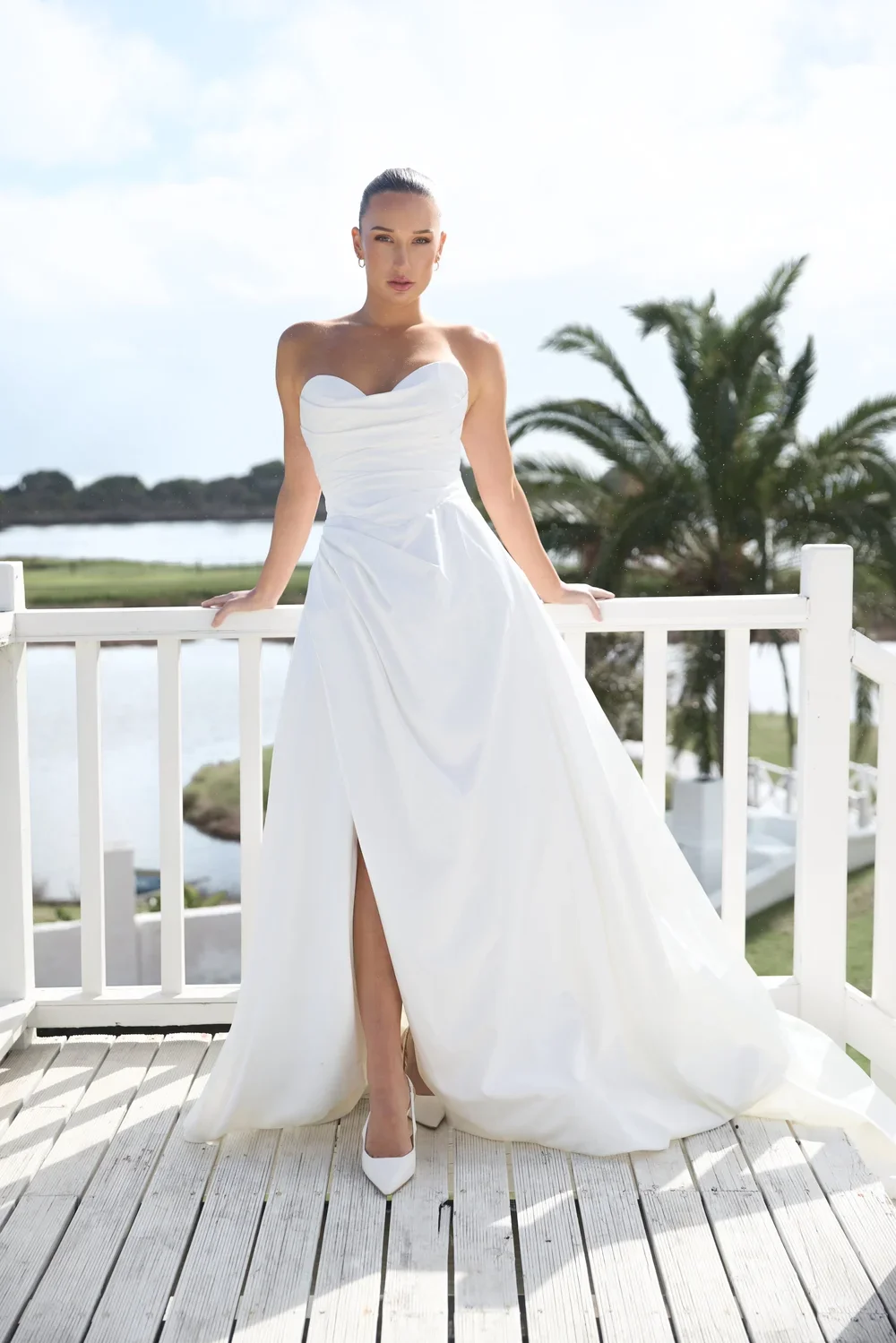A woman in a strapless Paige white wedding dress poses on a sunlit wooden balcony with palm trees and water in the background.