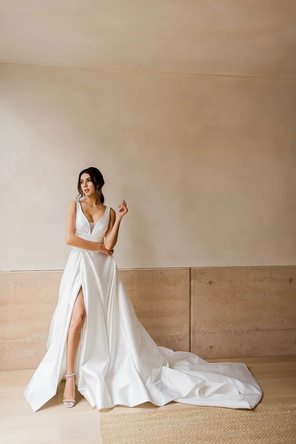 A woman in a white, floor-length wedding dress with a long train and a thigh-high slit stands indoors near a beige wall.