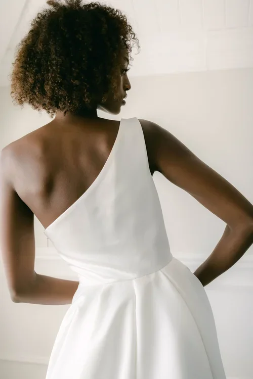 A woman with curly hair wears a one-shoulder EB22531-1Z sleeveless white dress, standing with her back turned in a softly lit room.