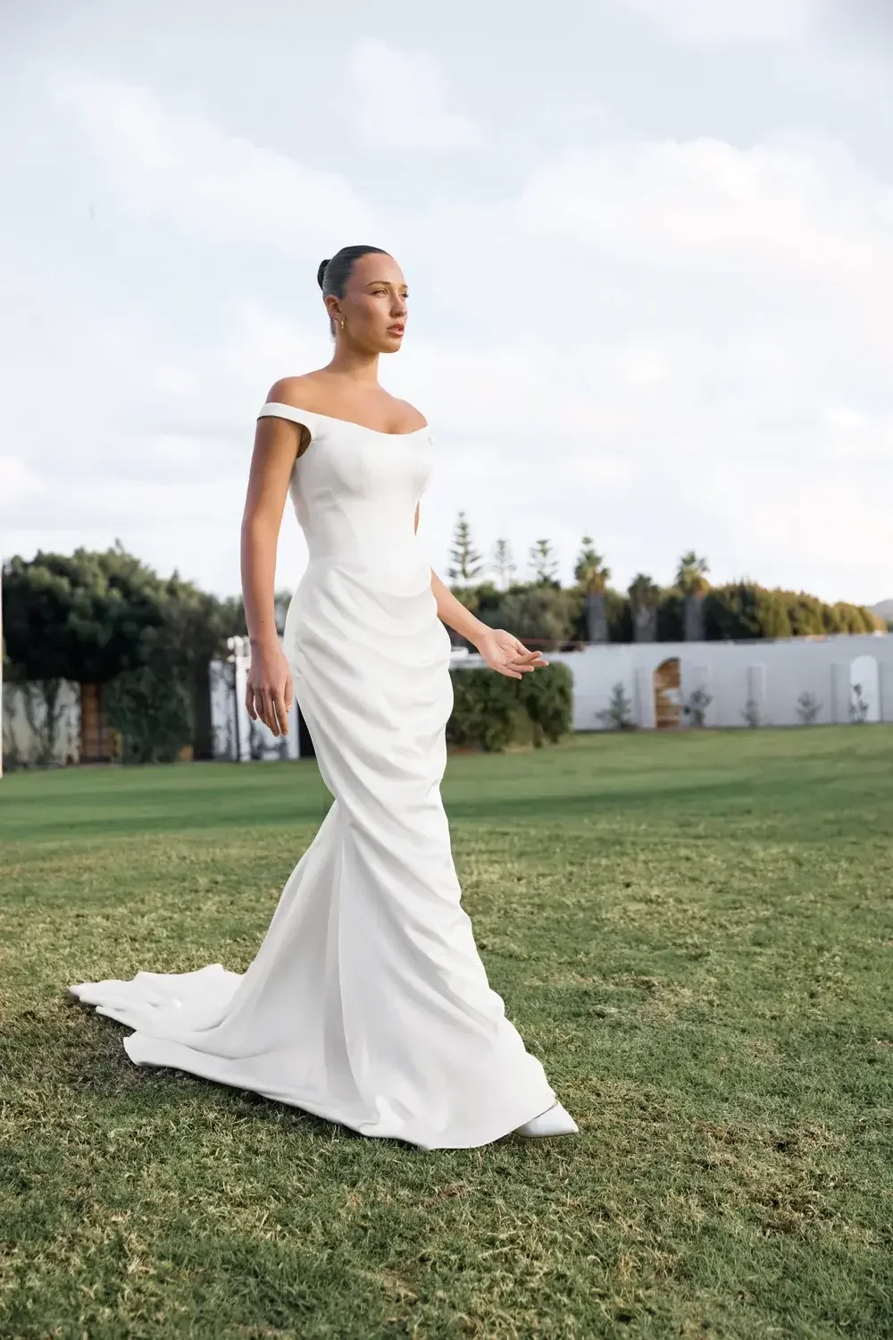 Model in an off-the-shoulder fitted white wedding dress with train walking on grass