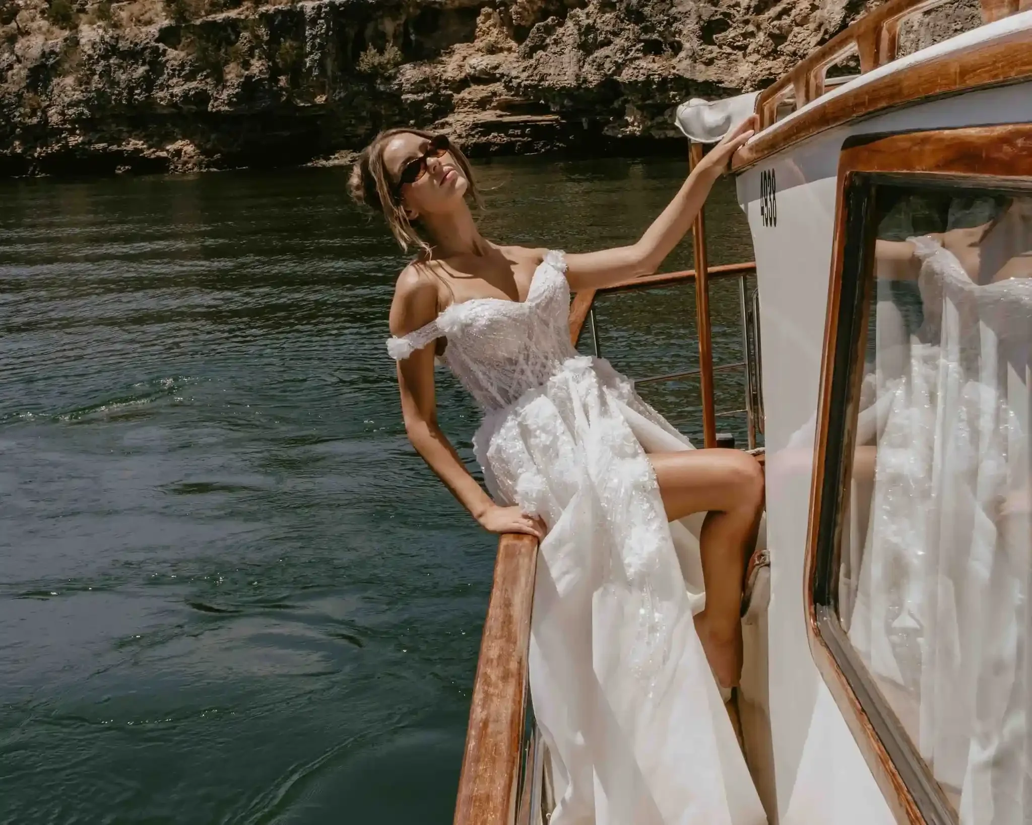 Woman in an off-shoulder white wedding dress and sunglasses on a boat by rocky cliffs