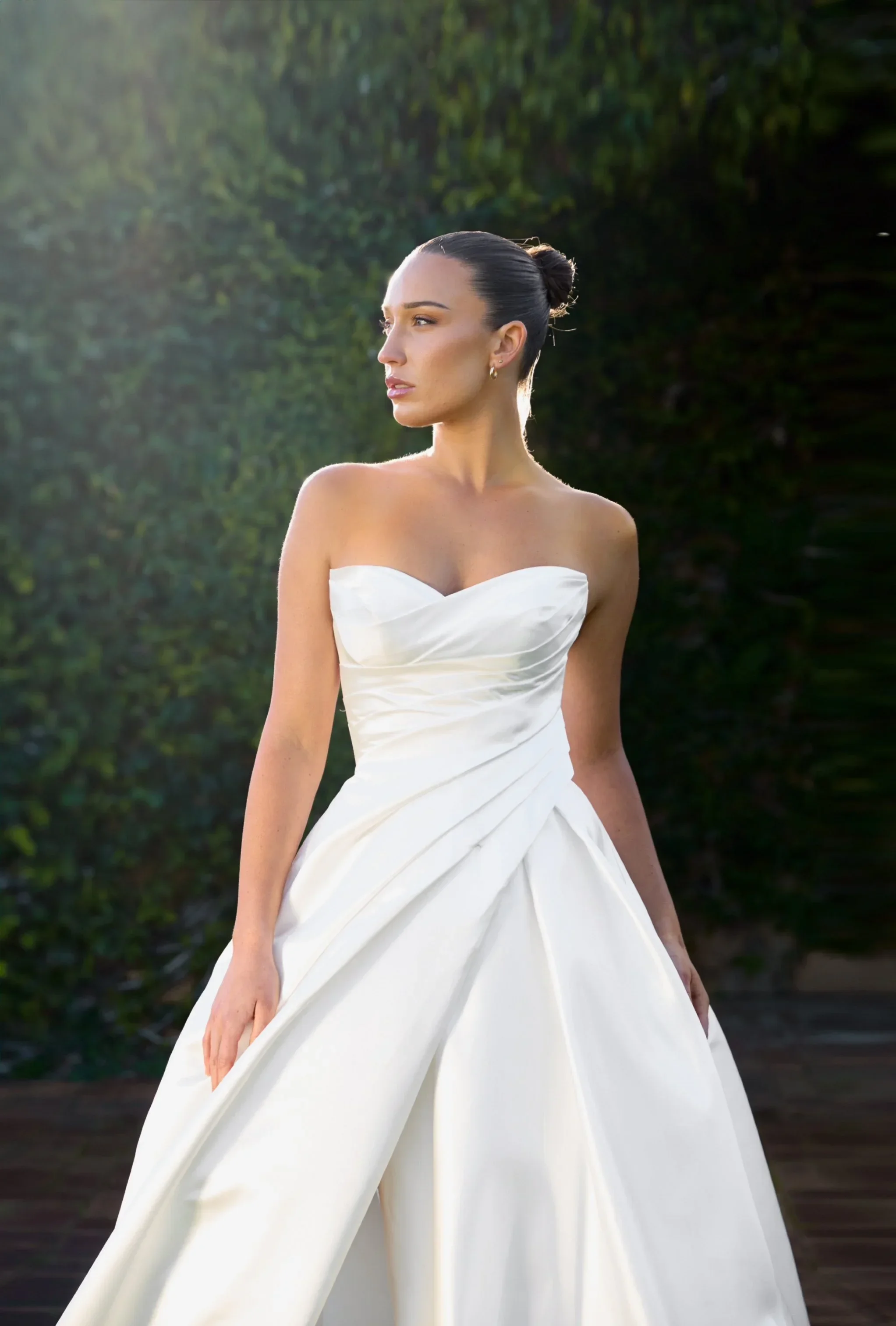 A woman in a Grace strapless white wedding dress stands outdoors, looking to the side. Her hair is styled in a sleek bun, and greenery is visible in the background. Soft sunlight shines on her from the top left.