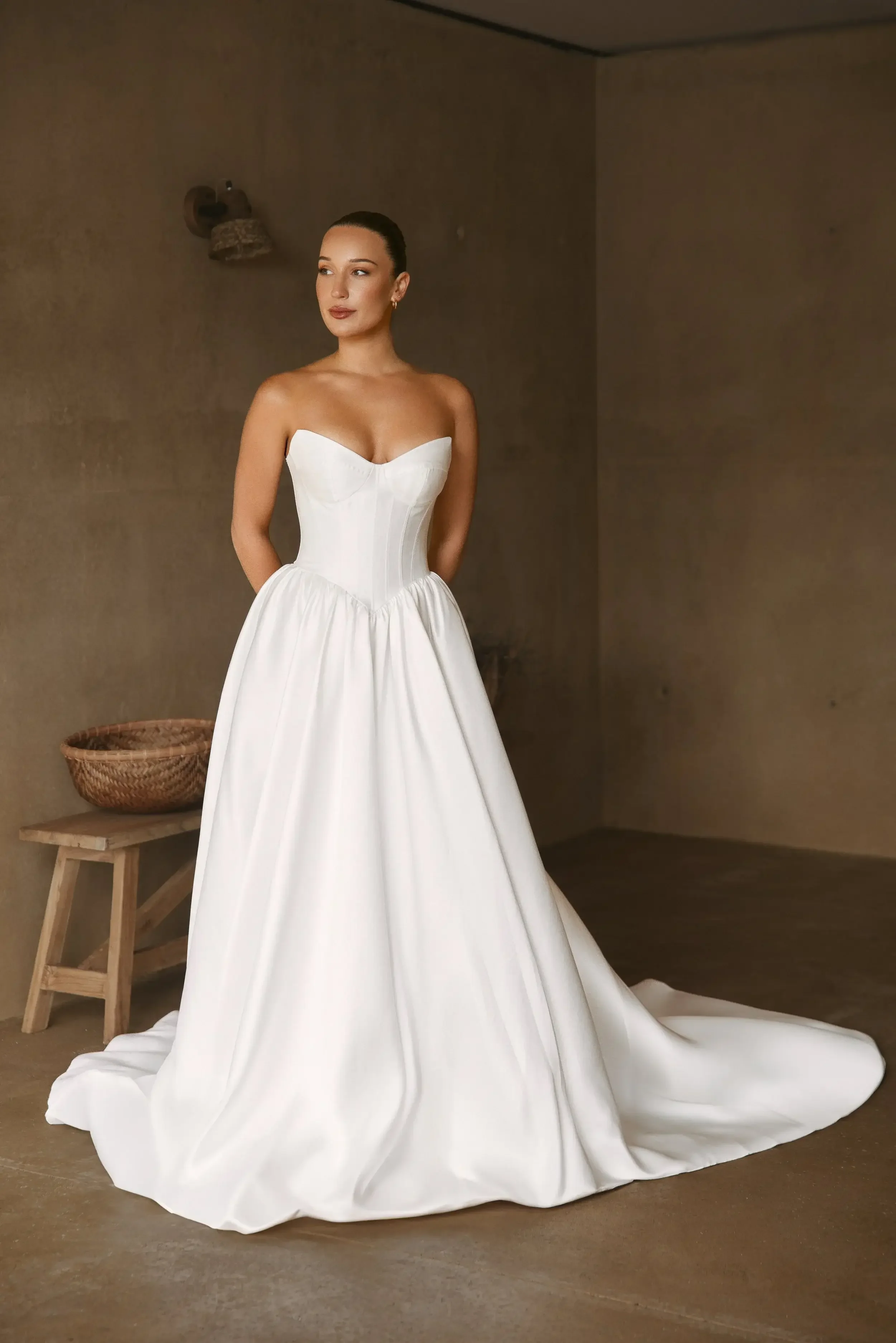 A woman in a Stella strapless white wedding gown with a fitted bodice and full skirt stands in a minimal, neutral-toned room with a basket on a wooden bench in the background.