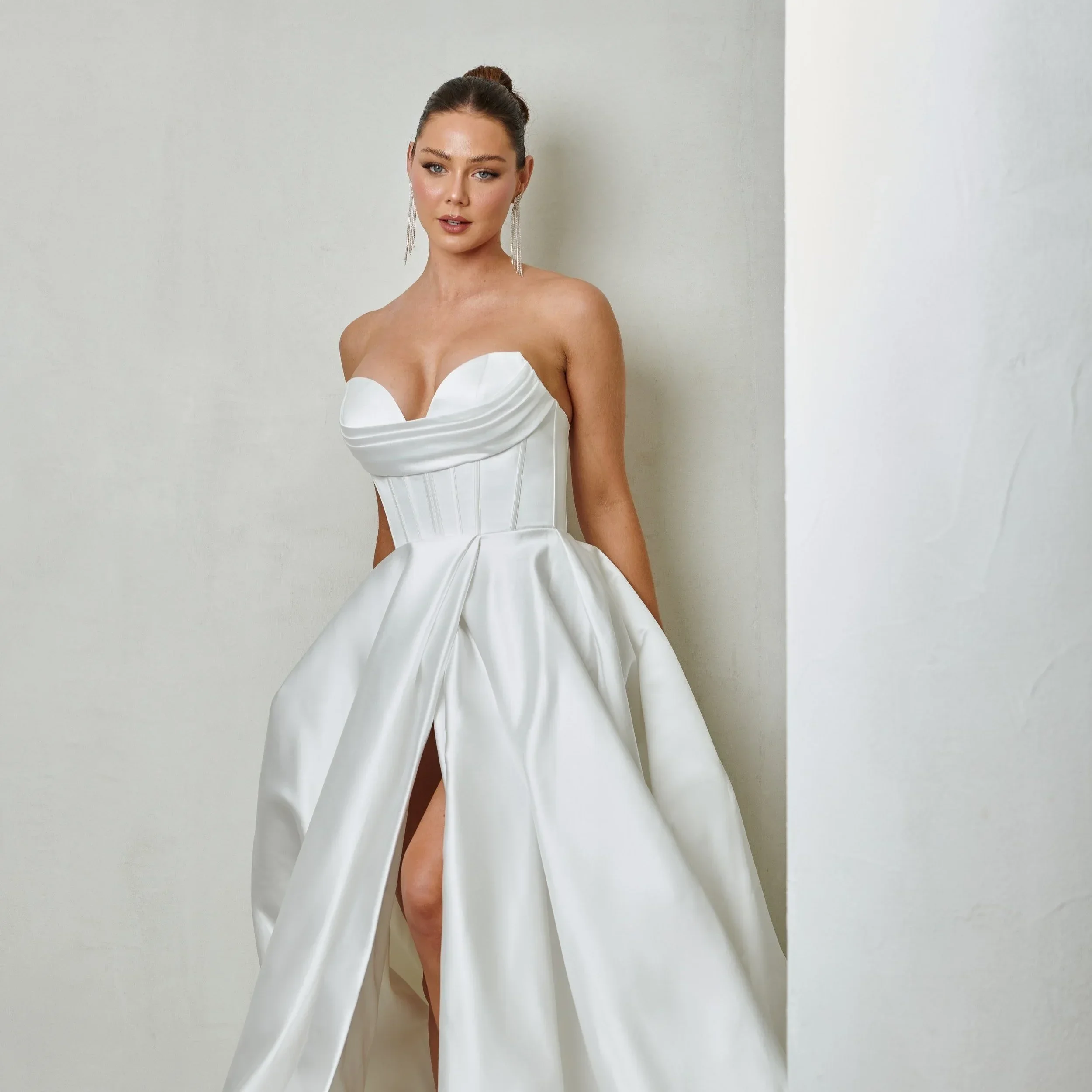 A woman in a strapless white wedding gown with a fitted bodice and a full skirt, standing against a plain white wall.
