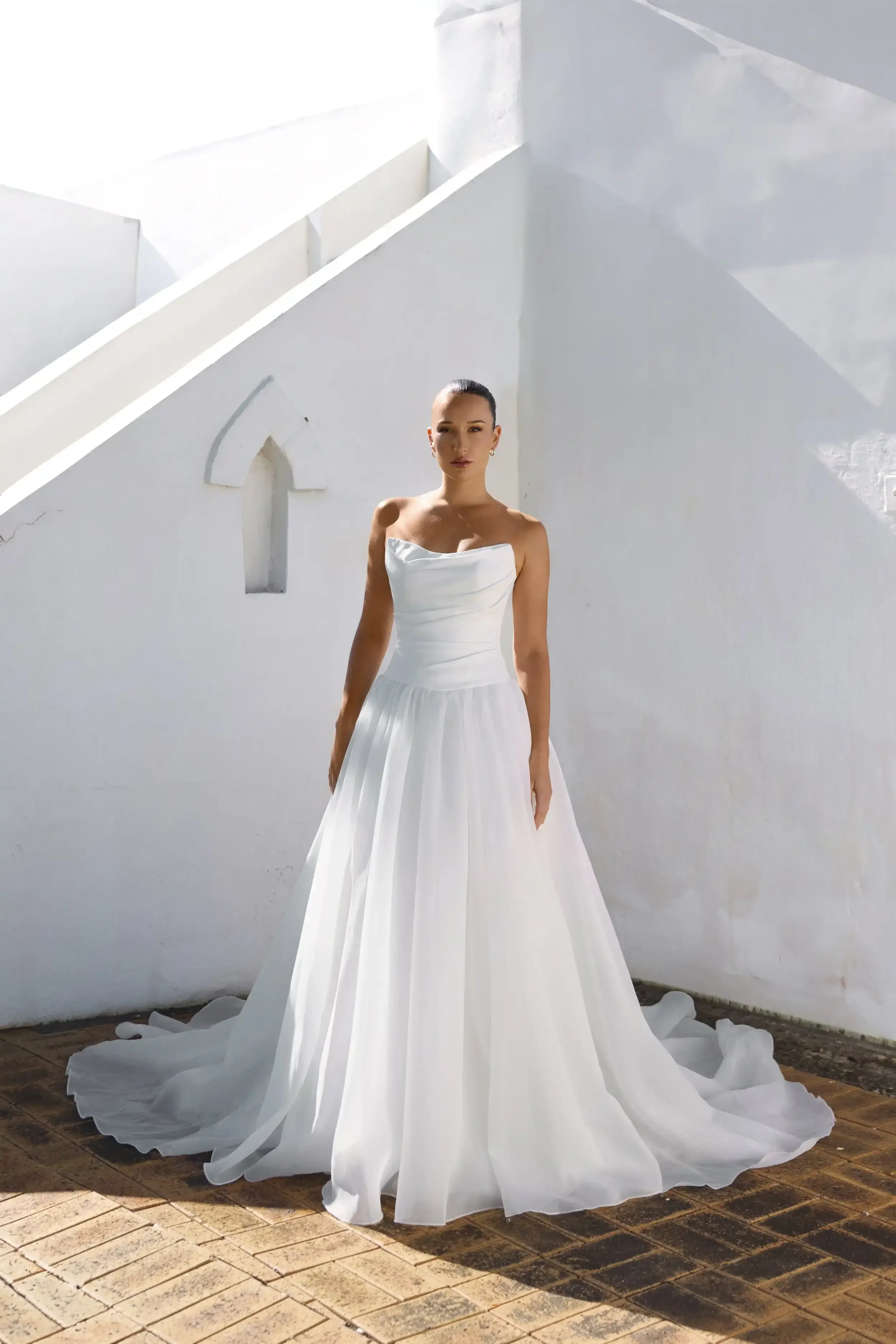 A woman in a Gigi white wedding gown with a full skirt stands on a brick floor against a sunlit white wall with geometric architectural features.