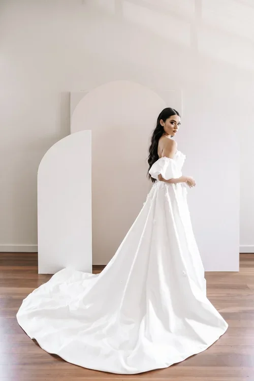 A woman in an off-the-shoulder white EB8614 wedding gown with a long train stands on a wooden floor in front of minimalist white, arched backdrops, looking over her shoulder.