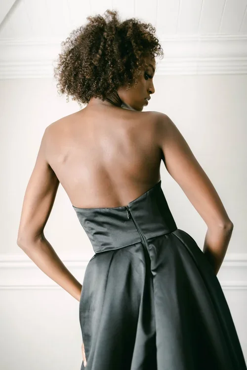 A woman stands in a bright, white room wearing a EB22456-1Z strapless, floor-length black gown with a full skirt and a slight train, her hair styled in natural curls.