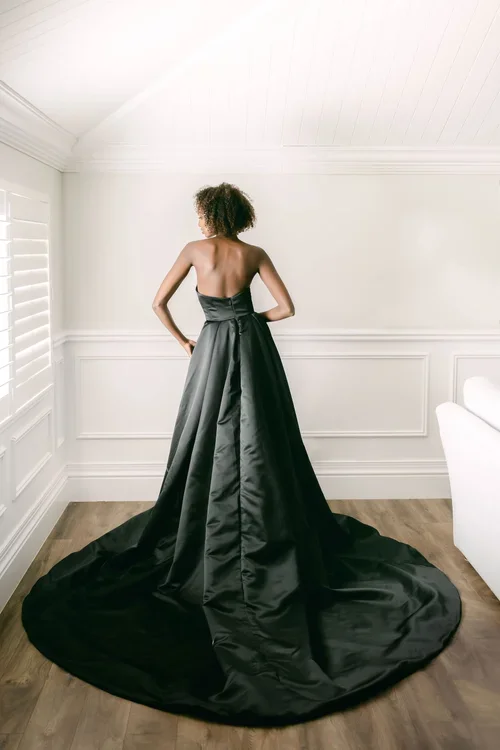 A woman stands with her back to the camera wearing EB22456-1Z strapless, floor-length black gown with a long train, in a bright, elegantly decorated room with white walls and wooden flooring.