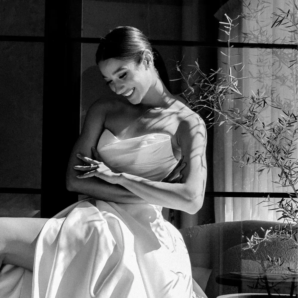 Model in a strapless Muse white dress seated on a sofa in soft sunlight, black and white