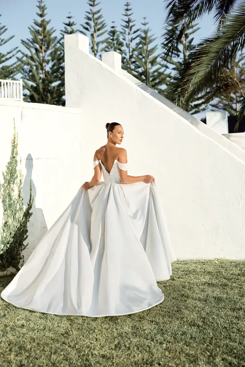 Back view of Elianna off-shoulder white bridal gown with flowing skirt outdoors